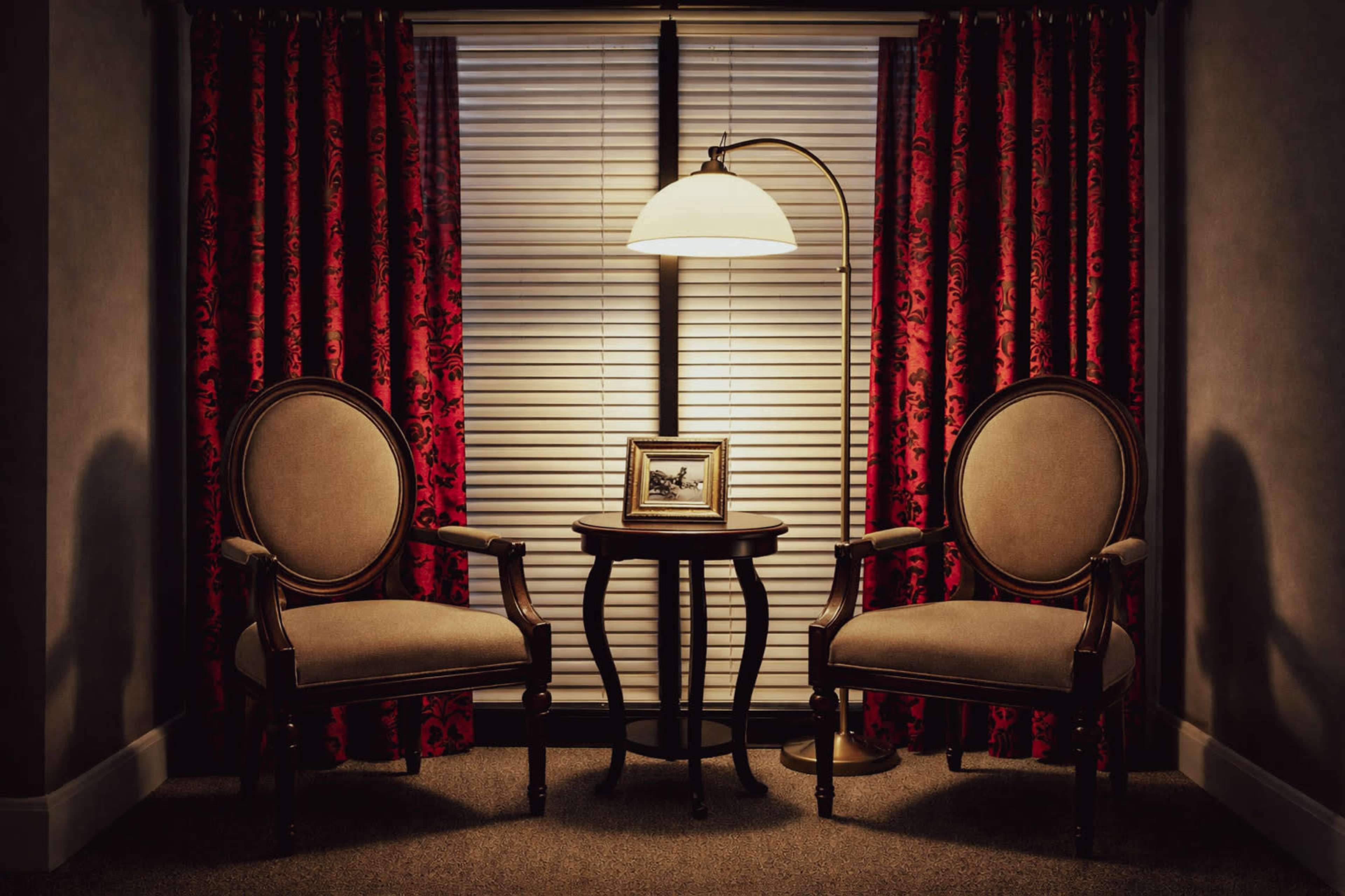 A cozy corner features two upholstered chairs flanking a small table with a framed picture, illuminated by a standing lamp, and surrounded by red patterned curtains.