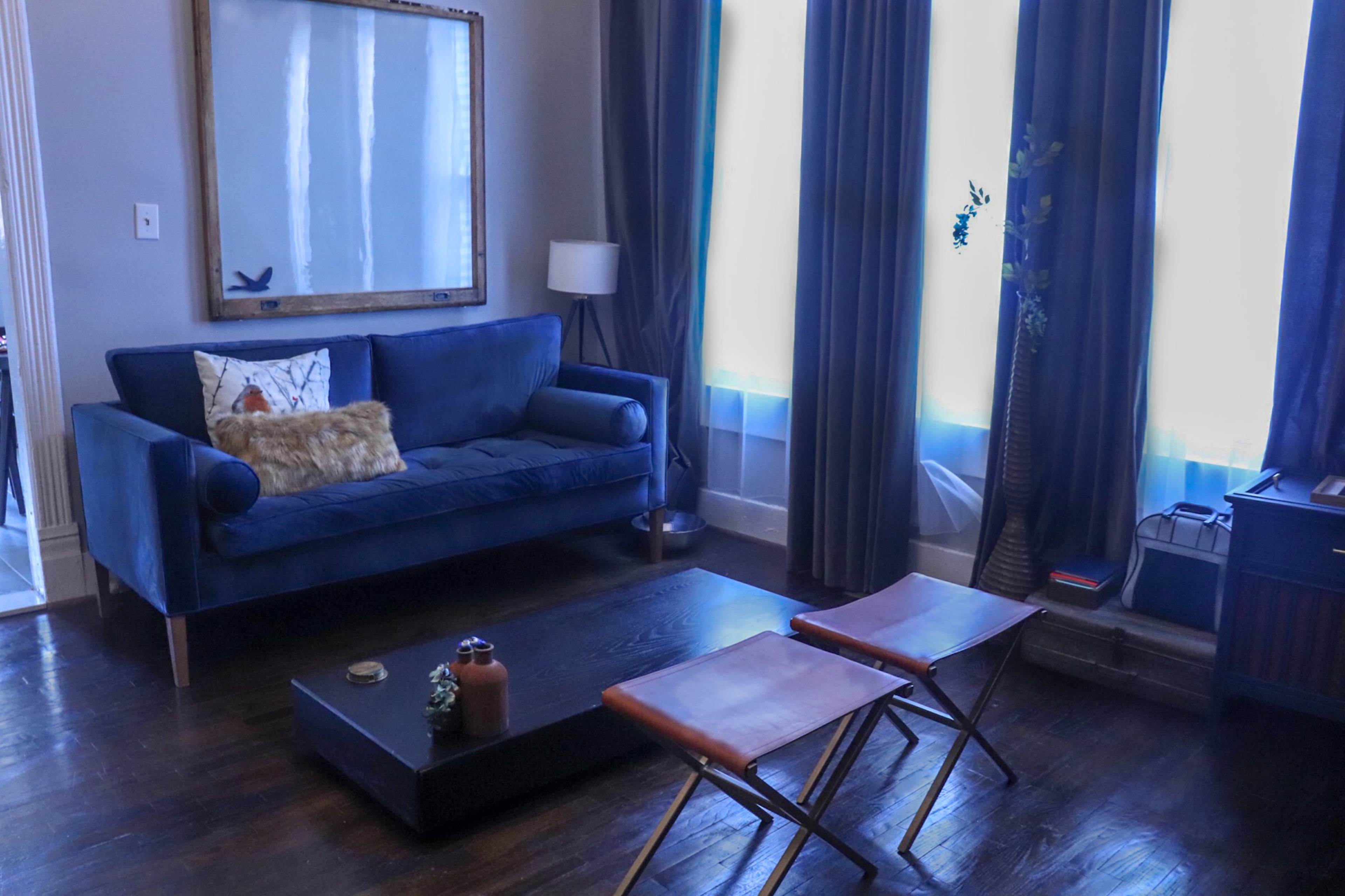 A living room with a blue sofa, a coffee table, and two folding chairs, illuminated by natural light from large windows.