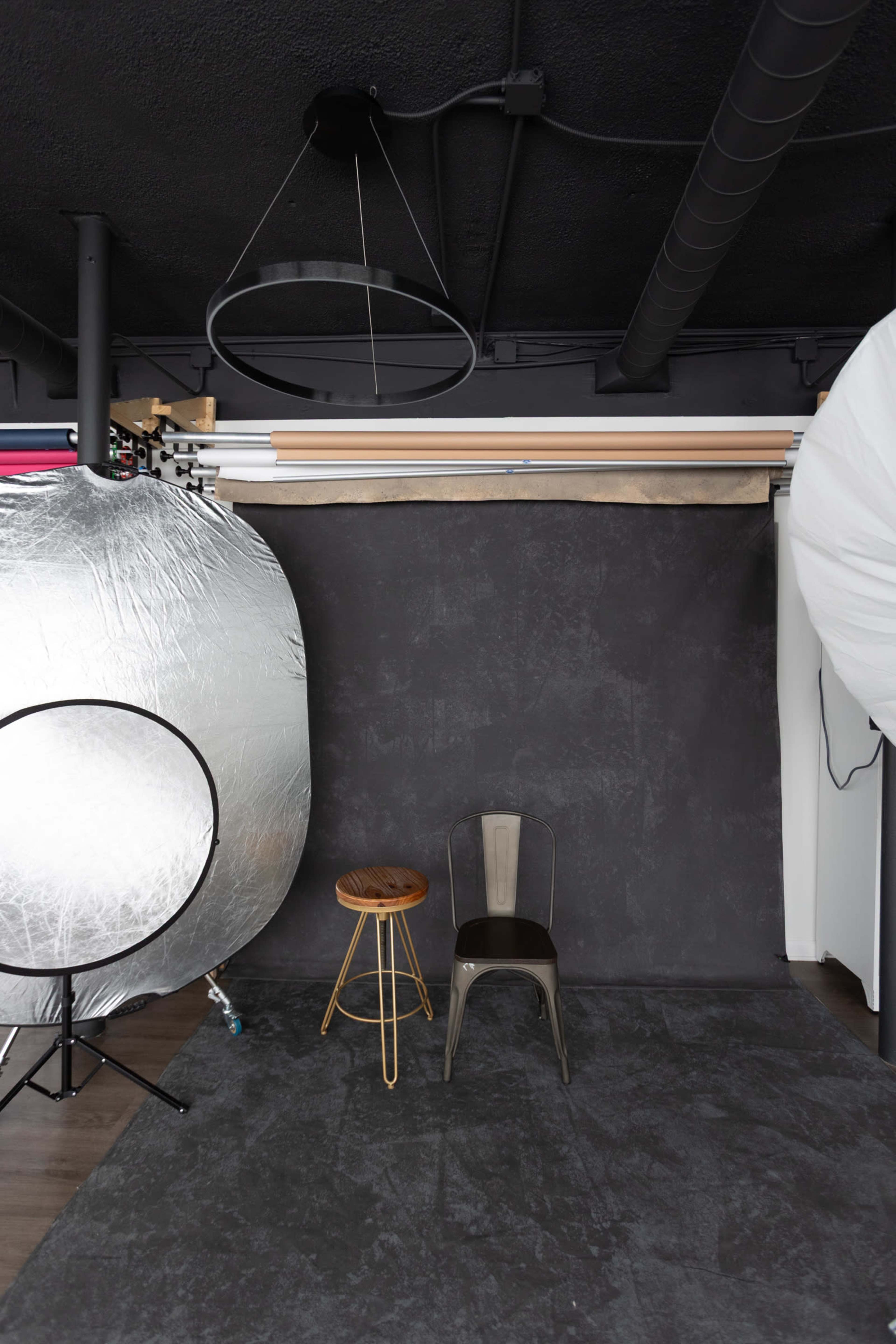The image shows a photography studio with a gray backdrop, a wooden stool, a metal chair, and various lighting equipment.