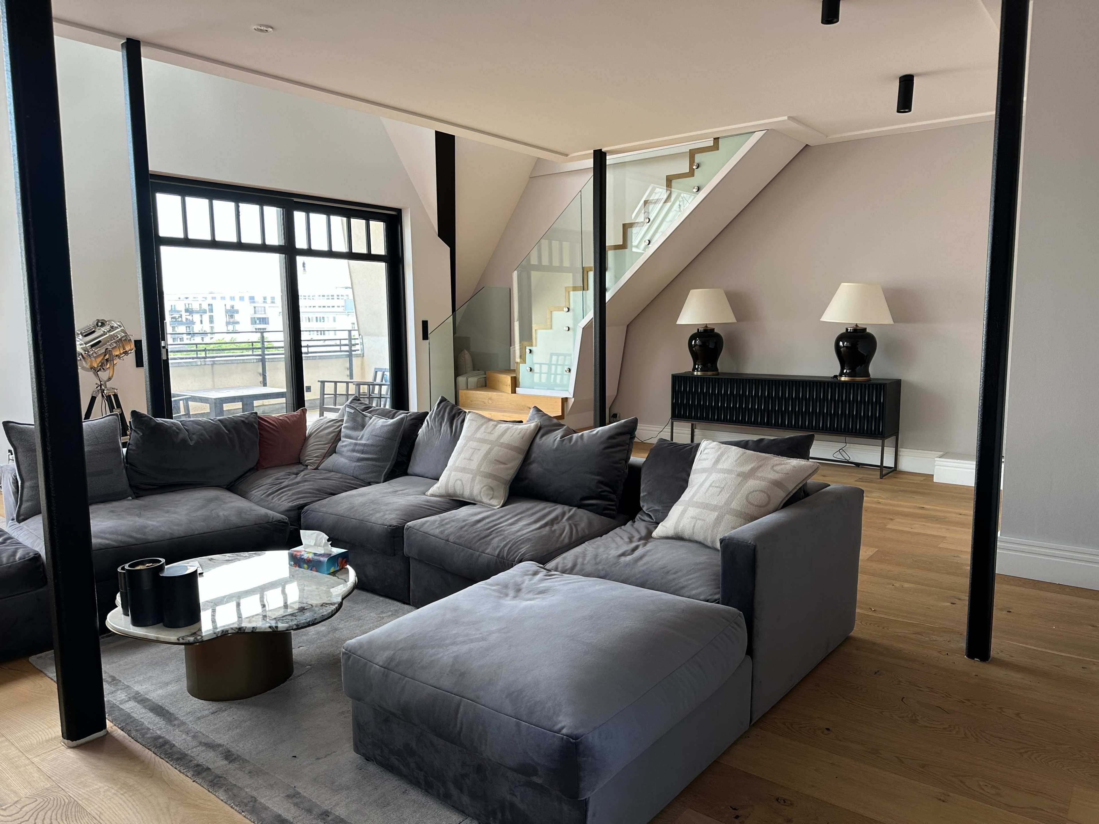 The image shows a modern living room with a large, grey sectional sofa, a glass coffee table, and a staircase in the background.