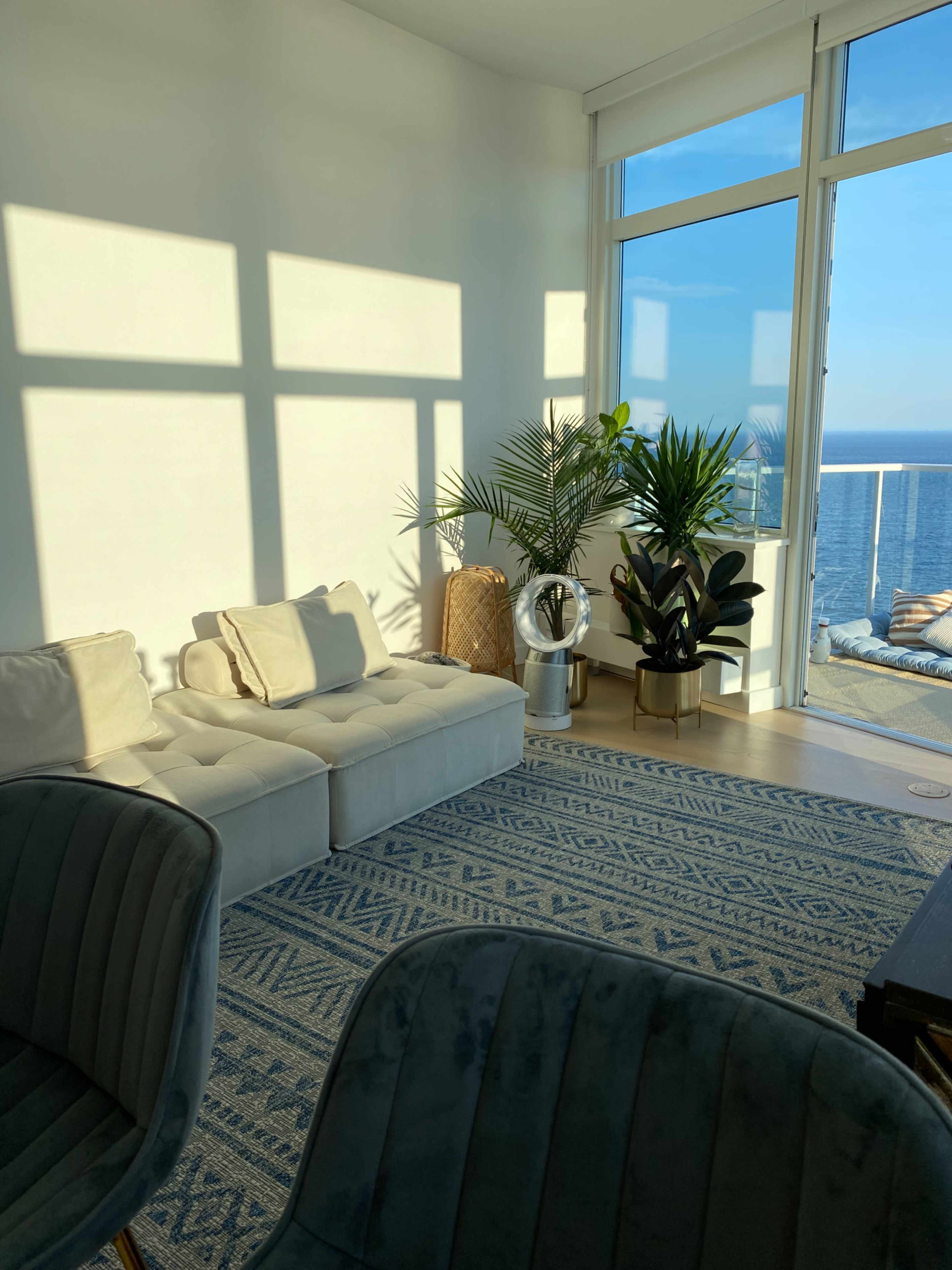 A modern living room features a sectional sofa, decorative plants, and large windows overlooking the ocean.