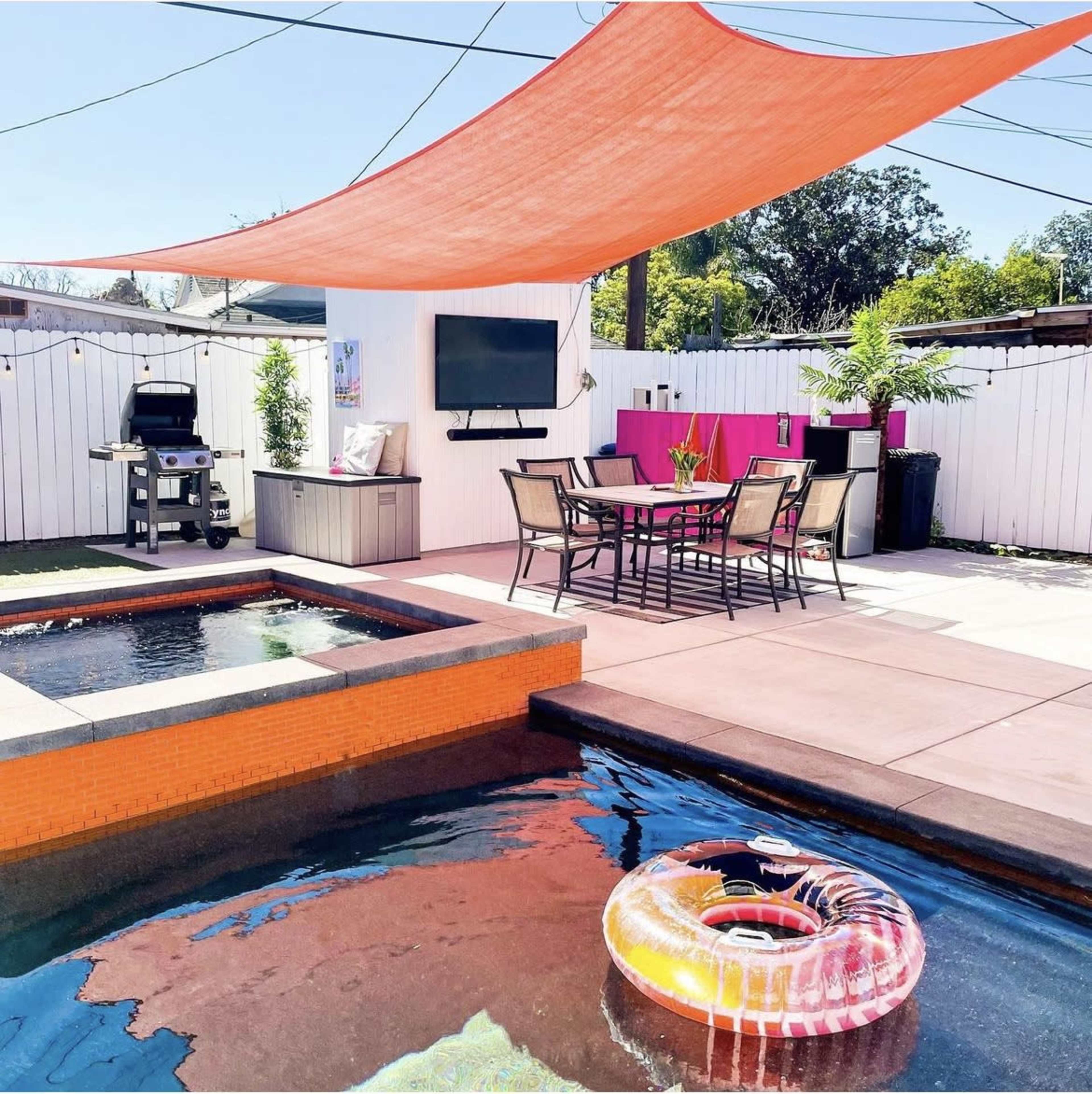 A backyard patio features a dining area with a table and chairs, a grill, a television mounted on the wall, and a swimming pool surrounded by a colorful floating ring and a fabric shade.