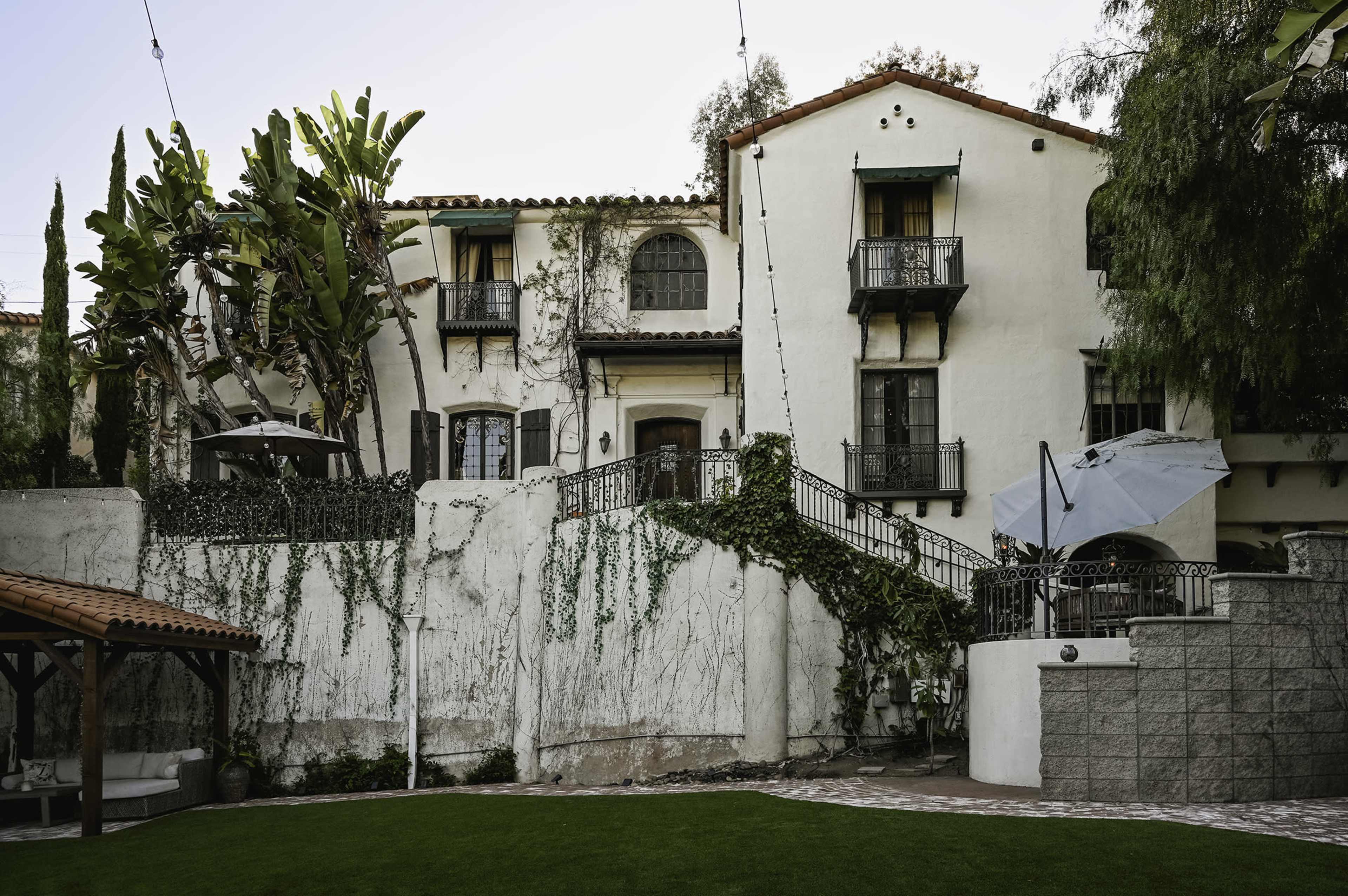 A multi-story Mediterranean-style house with balconies, surrounded by greenery and a landscaped yard.