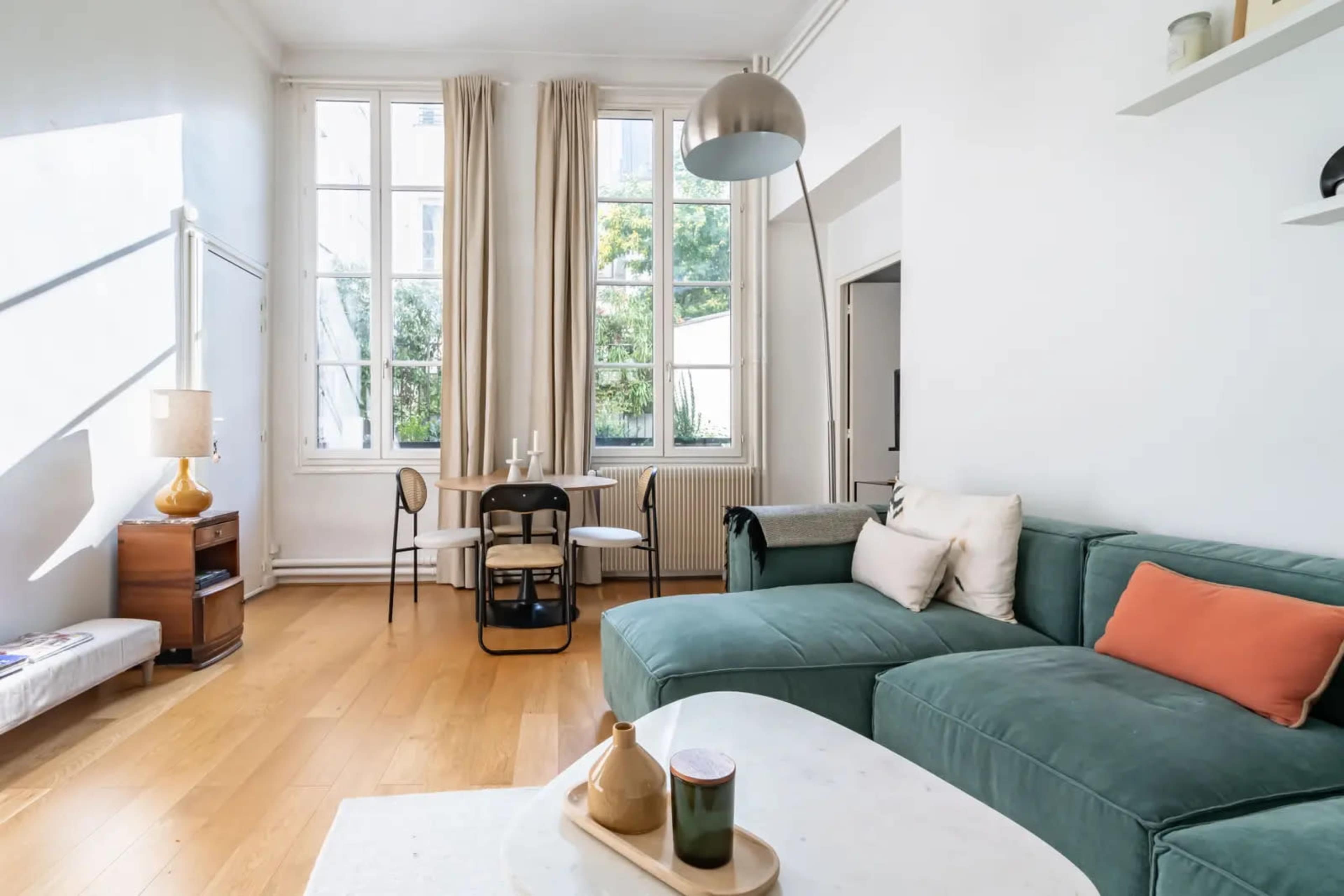 The image shows a bright, minimalist living space with a green sectional sofa, a wooden coffee table, and large windows letting in natural light.