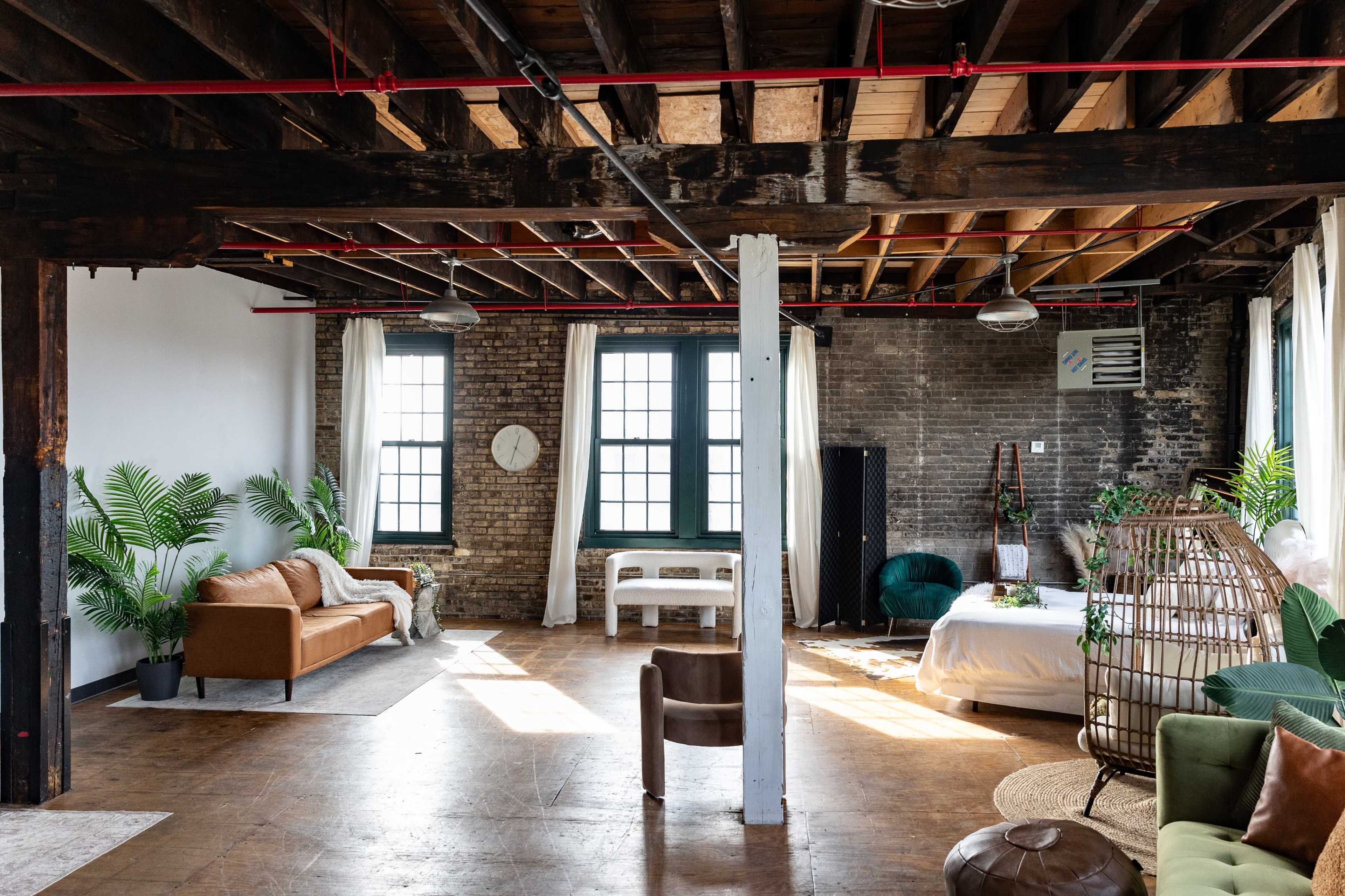 The image shows a spacious, industrial-style living area with exposed wooden beams, large windows, and a mix of plant decorations, furniture, and a bed in a well-lit setting.