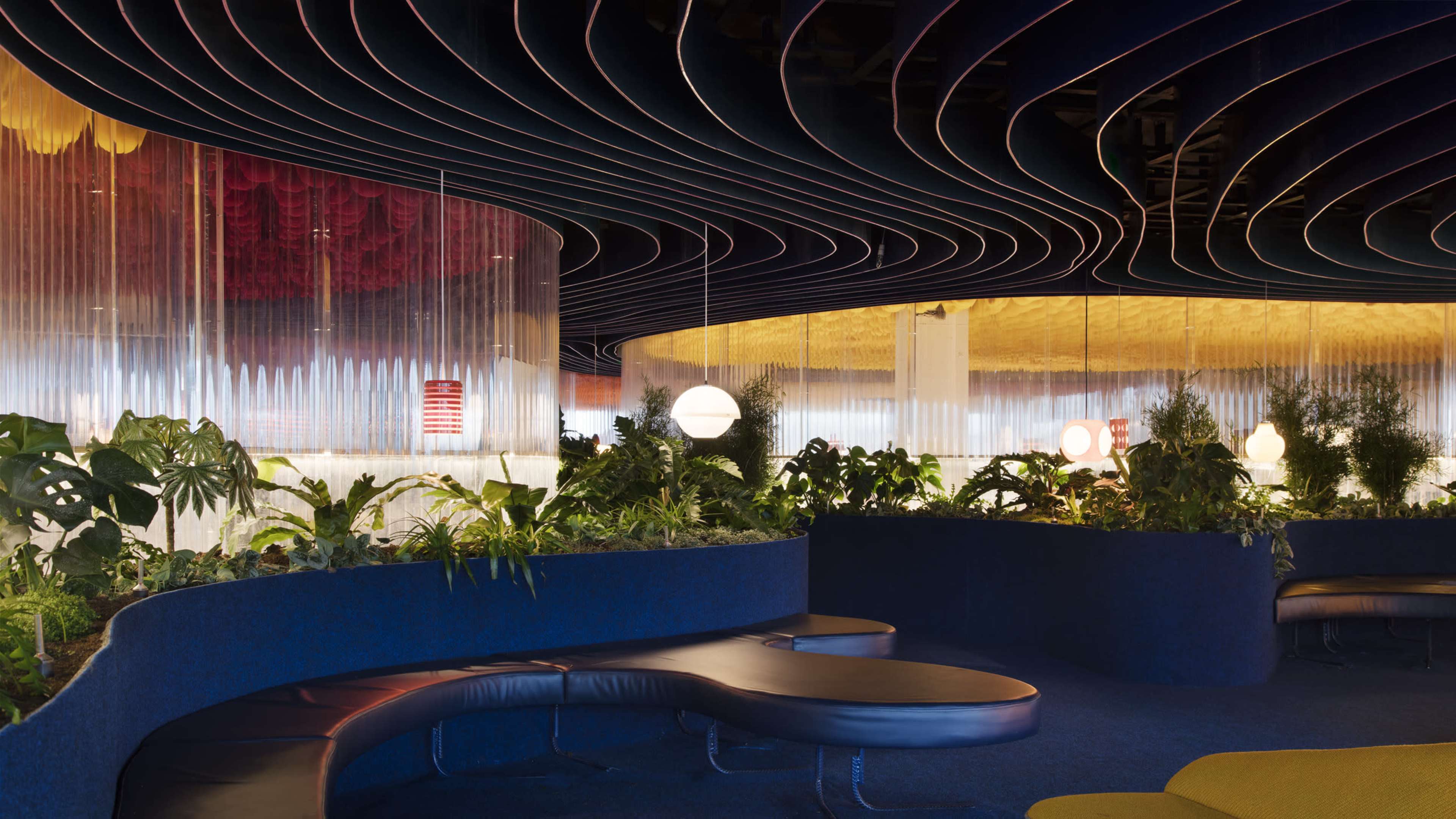 The image shows an interior space featuring curved ceiling patterns, vibrant lighting, and lush greenery interspersed among modern seating areas.