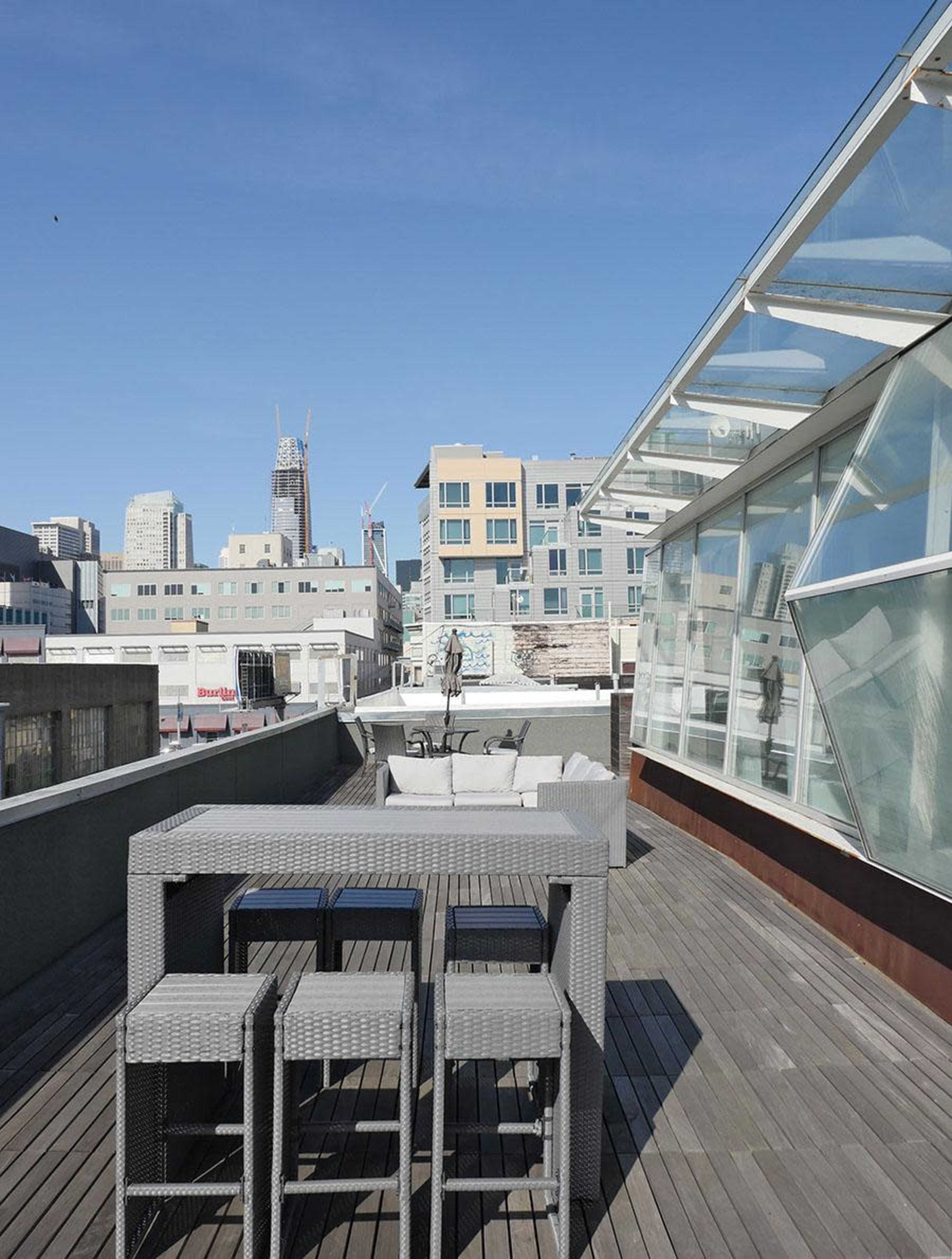 A rooftop patio with a wooden deck, featuring a wicker-style table and chairs, and a view of city buildings and a clear blue sky.