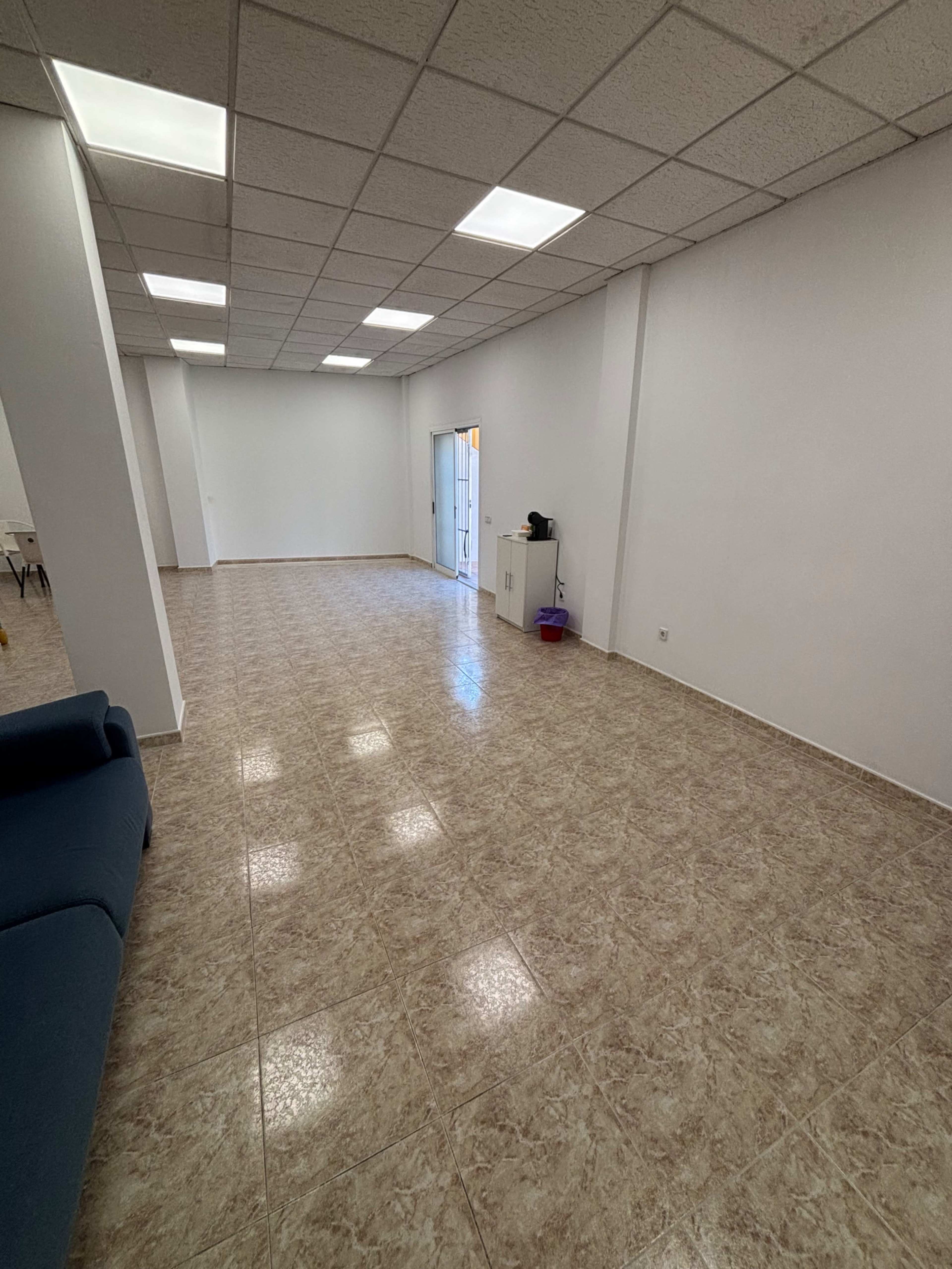 The image shows a spacious, empty room with beige tiled flooring and white walls, featuring a few ceiling lights and a door leading outside.