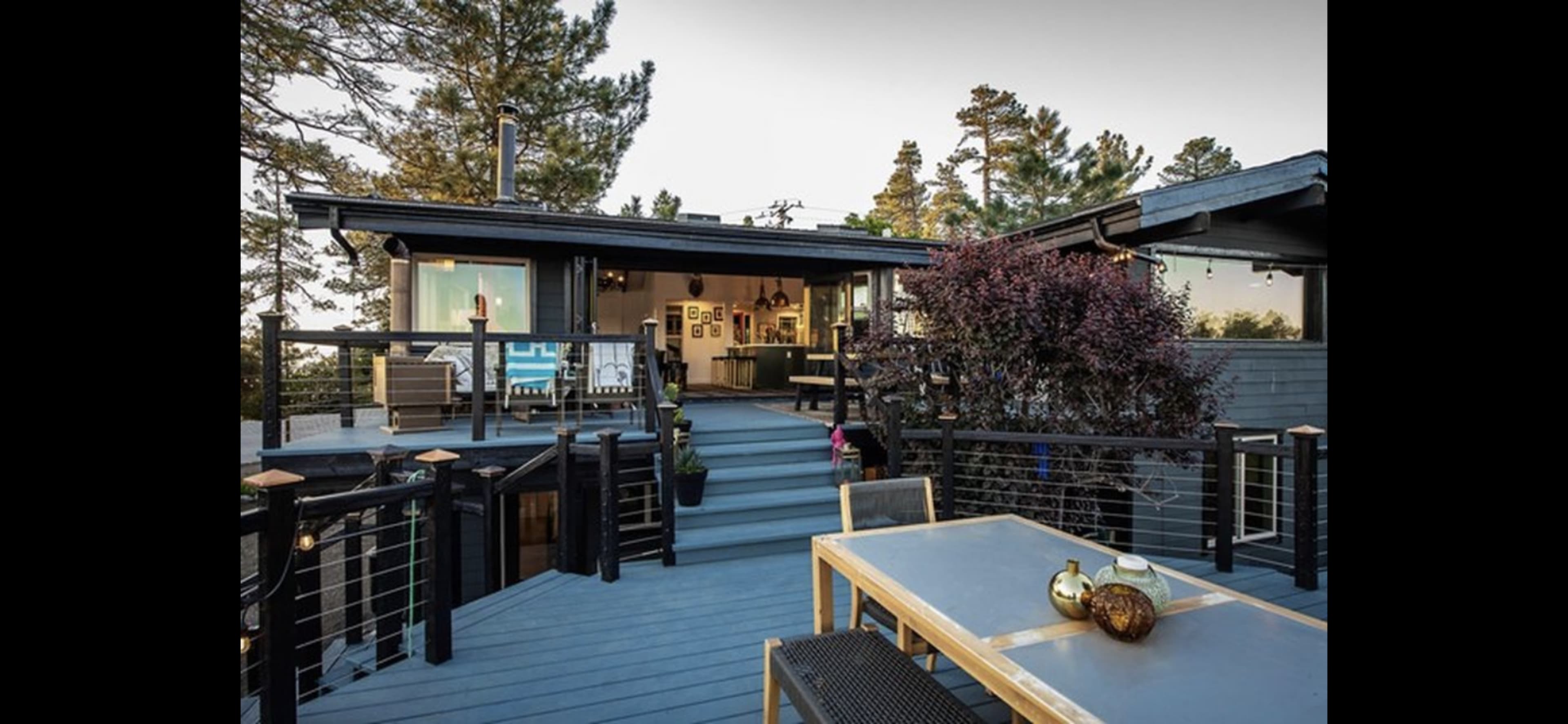 The image shows a modern wooden deck with a dining table, leading to a contemporary house surrounded by trees.