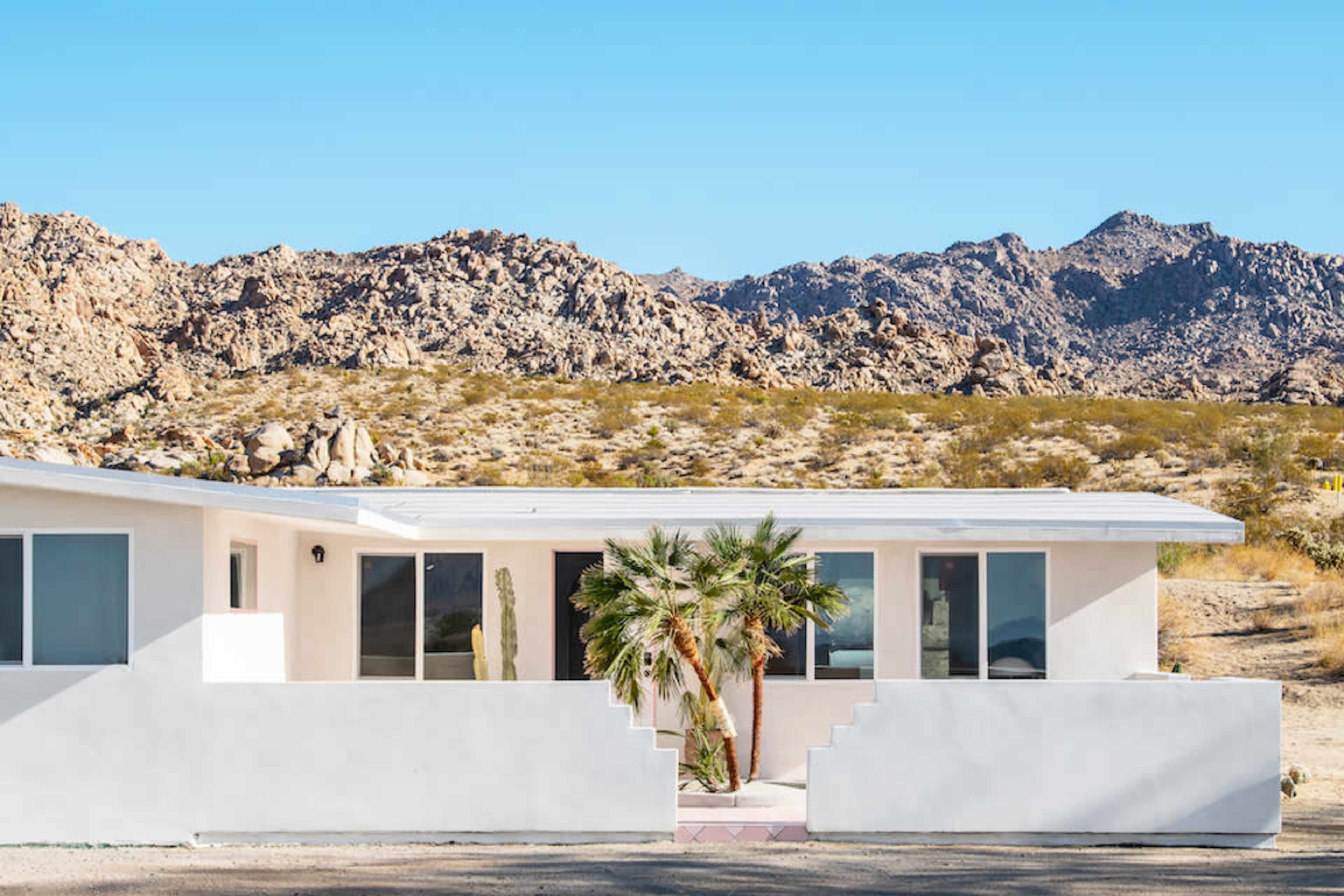 A modern white house with large windows and a palm tree is situated in a rocky desert landscape.