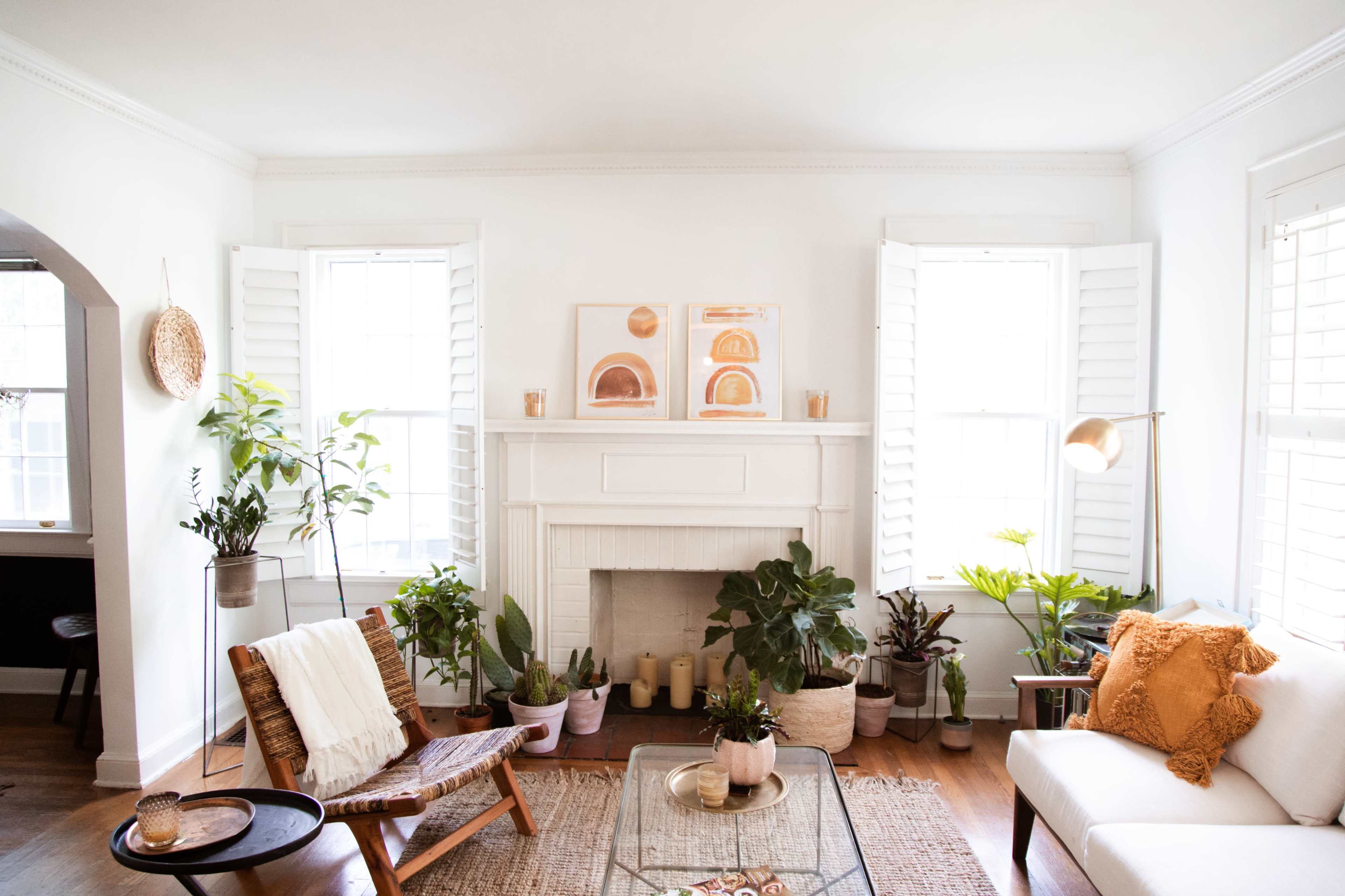 A cozy living room features a white wall, large windows, a fireplace with artwork above it, and various indoor plants arranged around a glass coffee table.