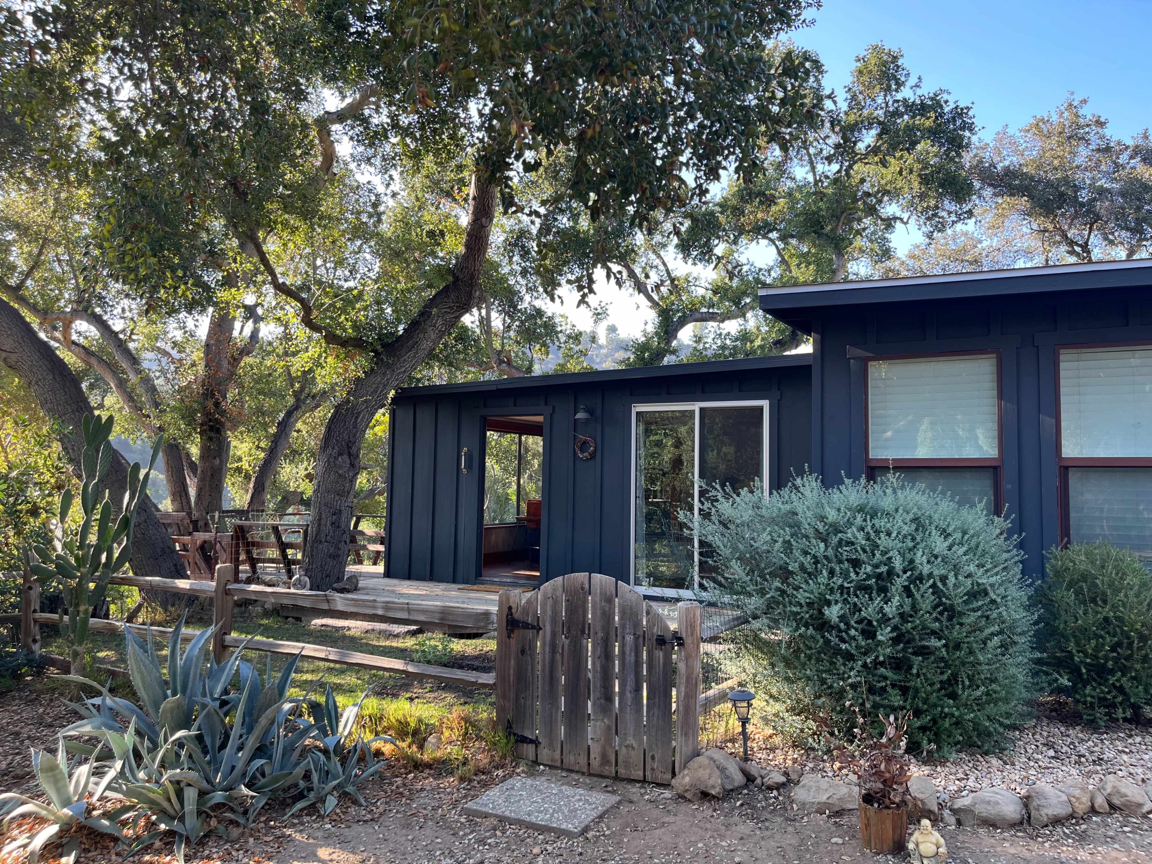 A modern, dark blue house with large windows is surrounded by trees and landscaping, featuring a wooden gate and a patio area.