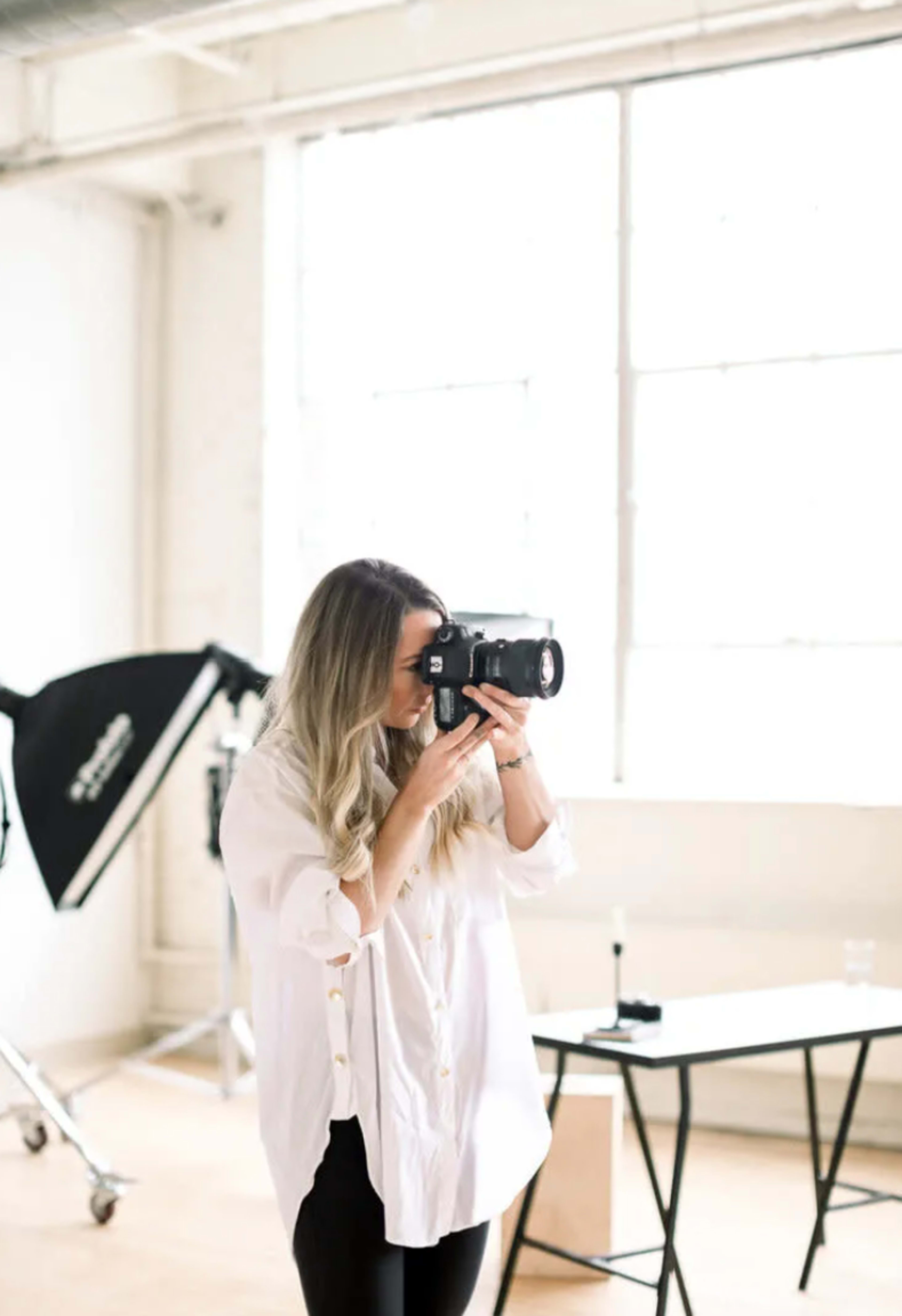 A woman is holding a camera and taking a photo in a bright, minimalist studio with large windows.