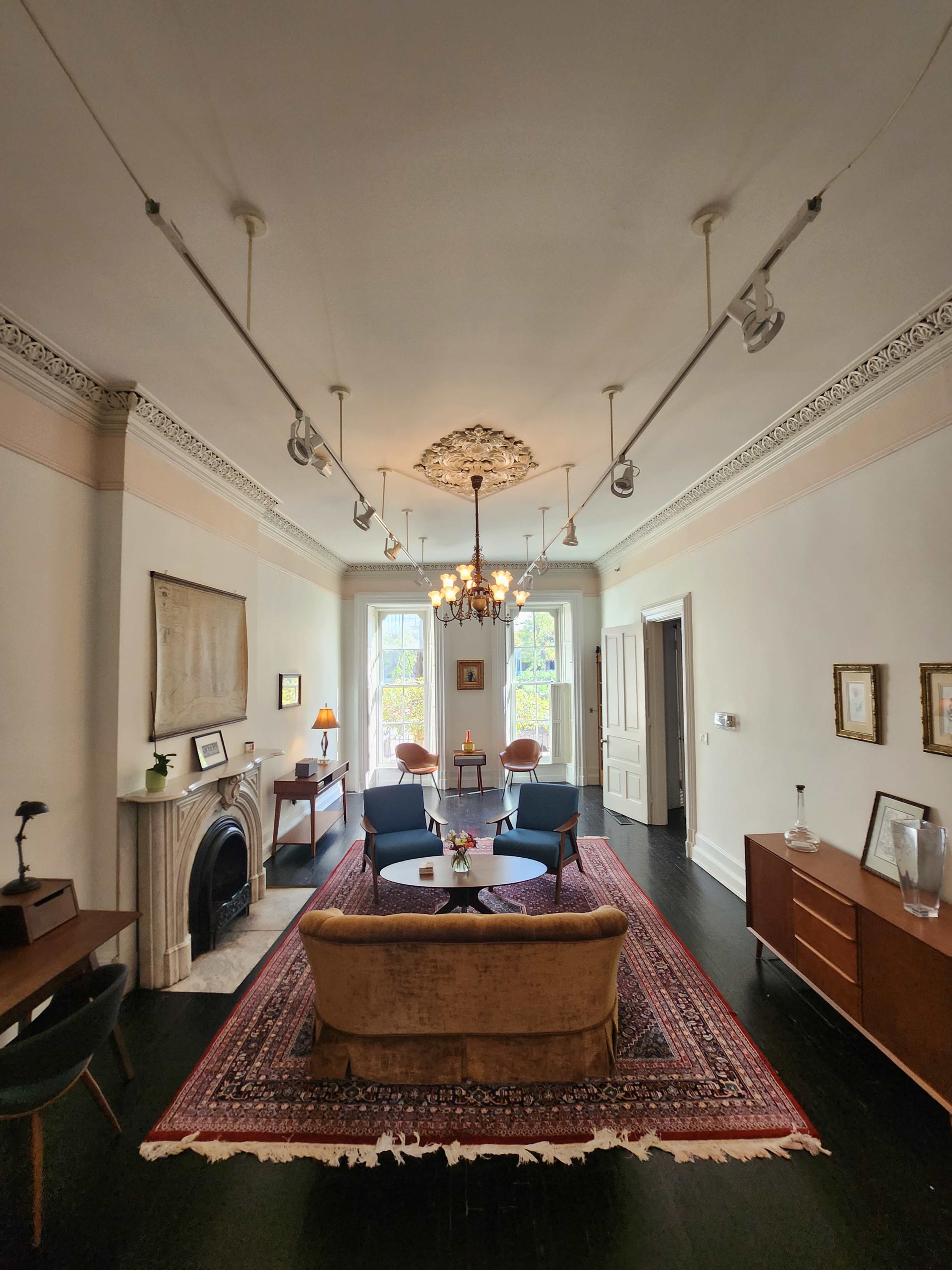 A spacious living room with a central chandelier, a patterned rug, a seating arrangement, and decorative elements on the walls.