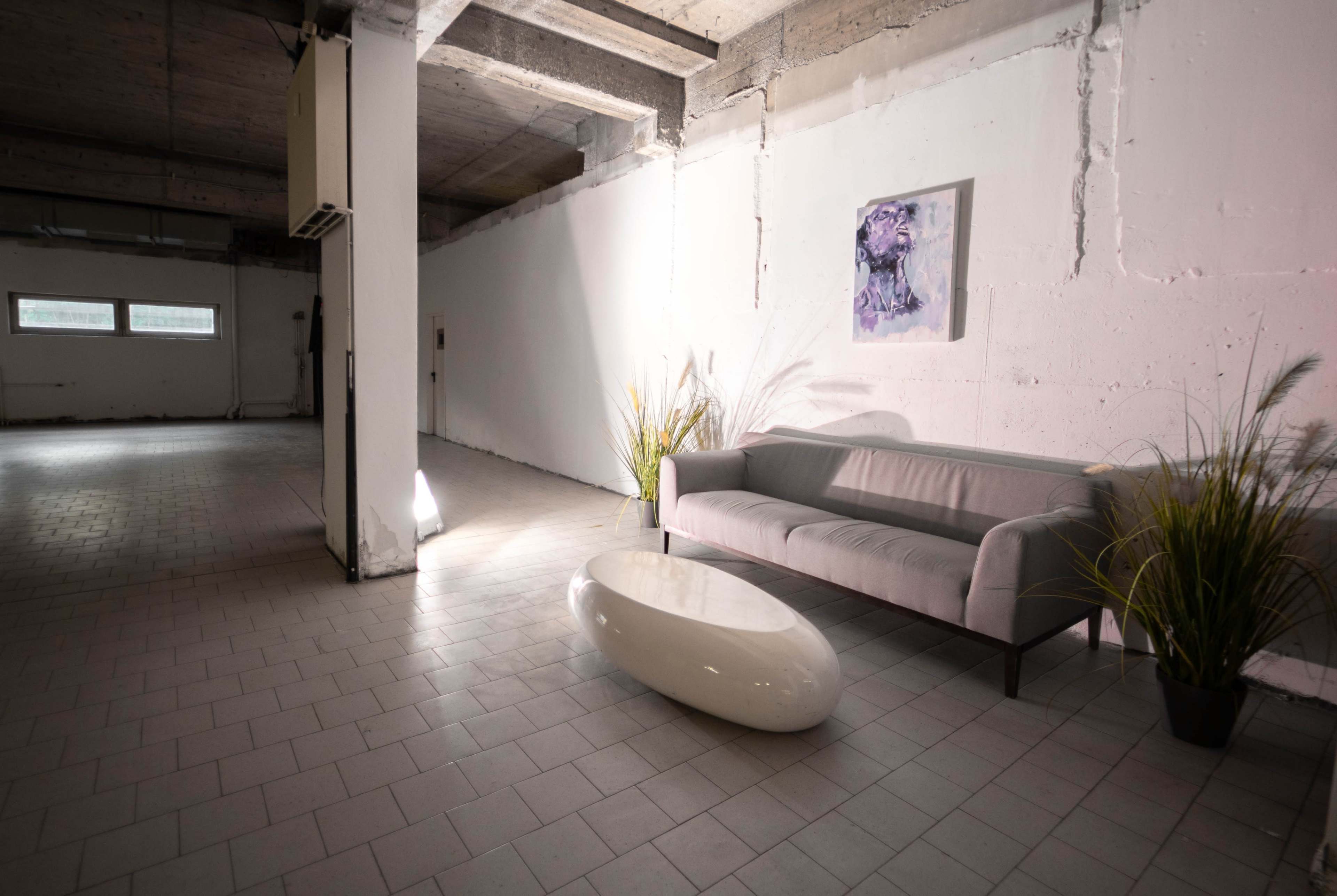 The image shows a minimalist interior space with a light gray couch, a round white coffee table, and a framed artwork on the wall, illuminated by natural light.