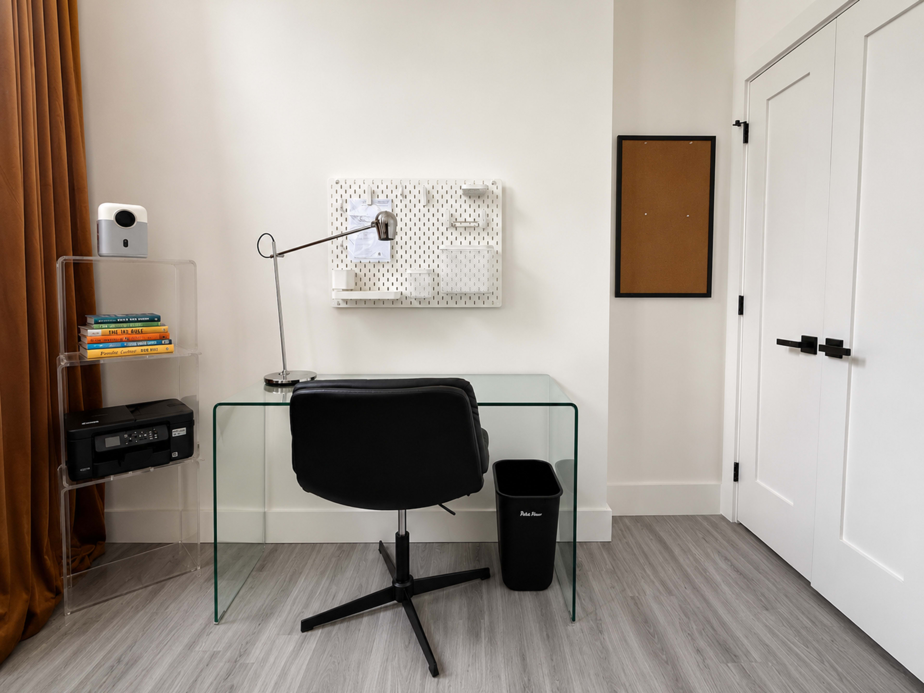 A modern workspace features a glass desk with a black chair, a wall-mounted organizer, a lamp, and a trash bin against a light-colored wall.