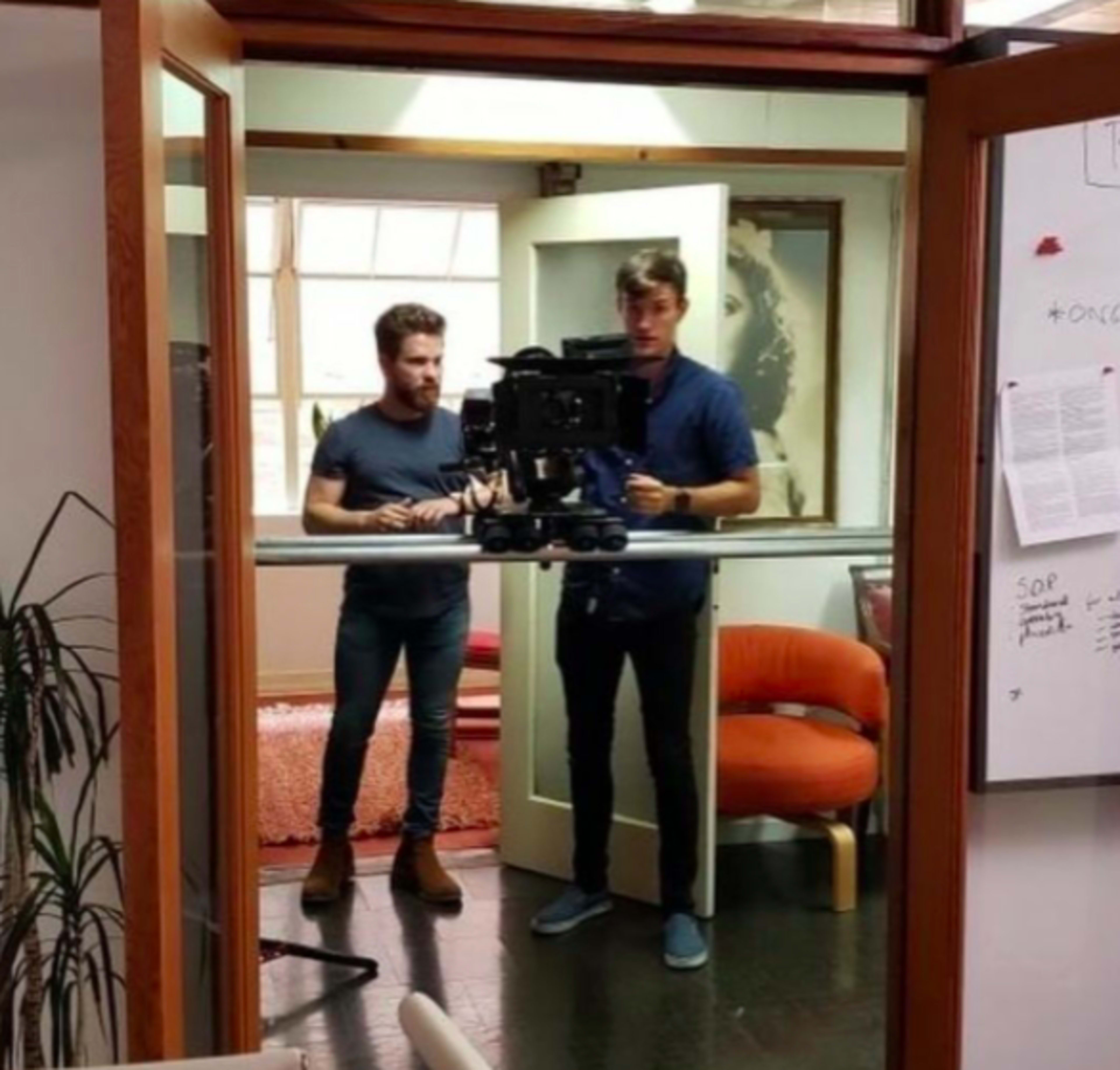 Two men are filming in an office setting, positioned in front of a camera mounted on a tripod.