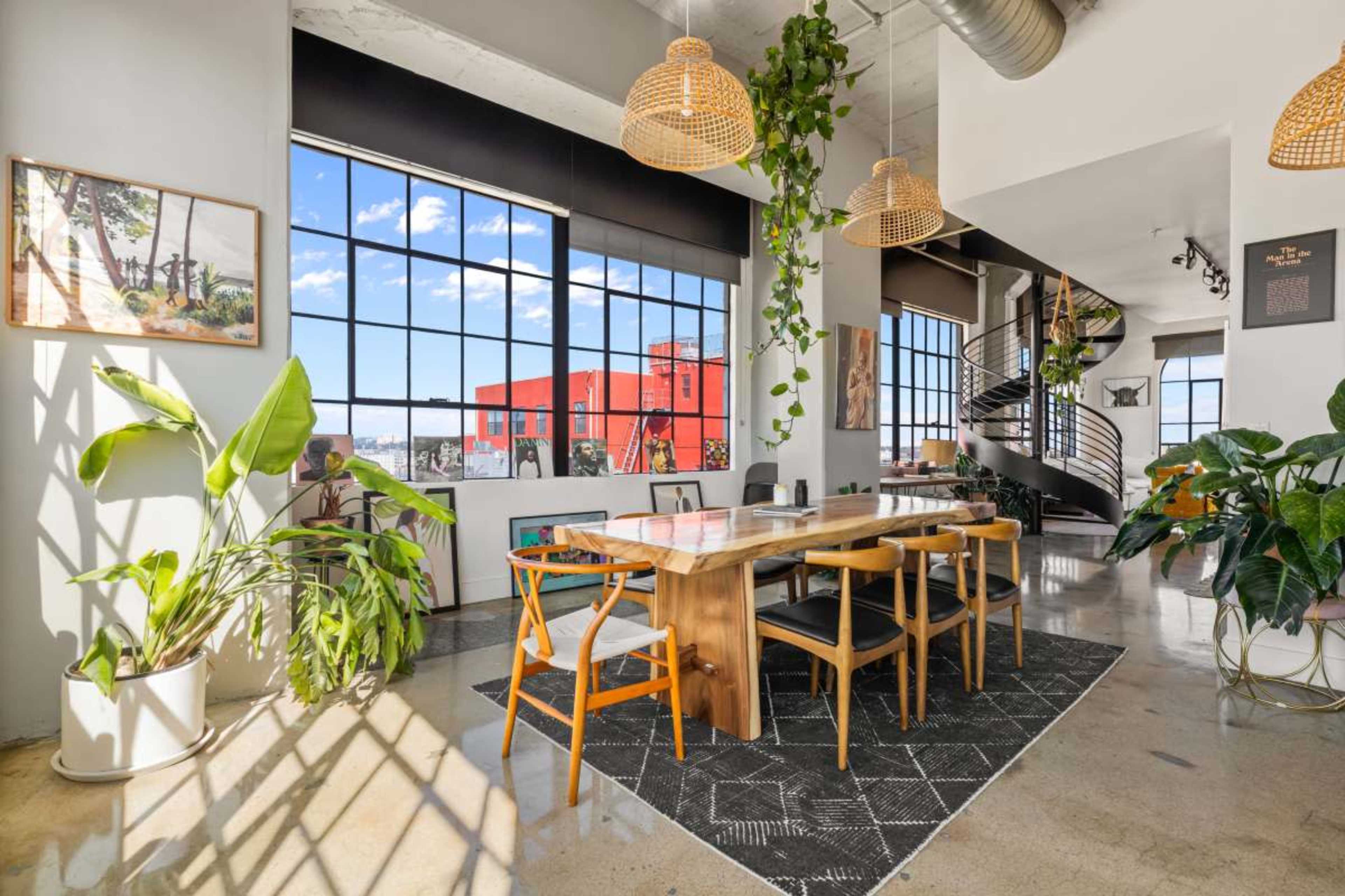 The image showcases a modern dining area with a wooden table surrounded by chairs, large windows offering a view of the outdoors, and decorative plants enhancing the space.