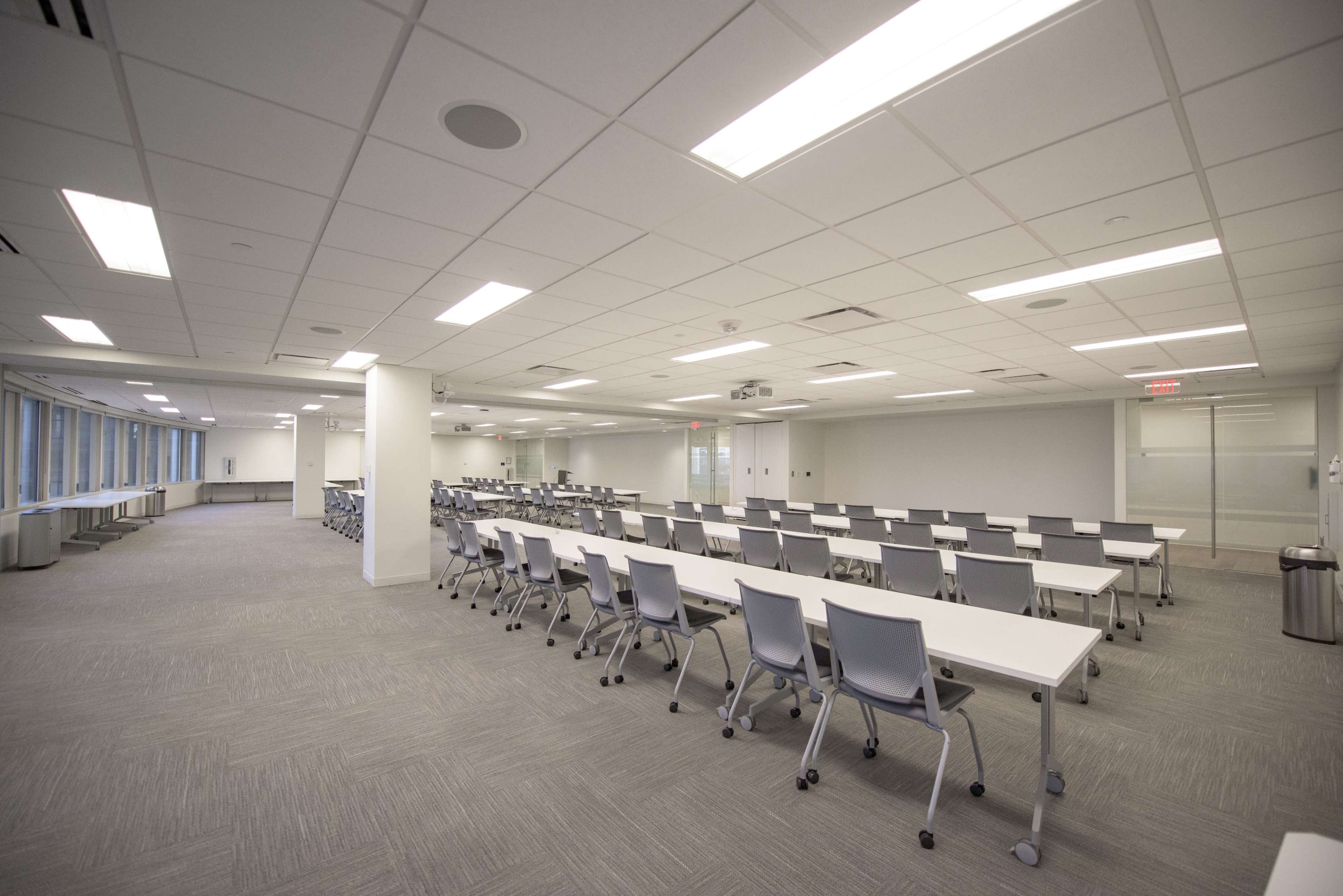 The image shows a spacious, modern conference room with multiple rows of white tables and gray chairs arranged neatly.
