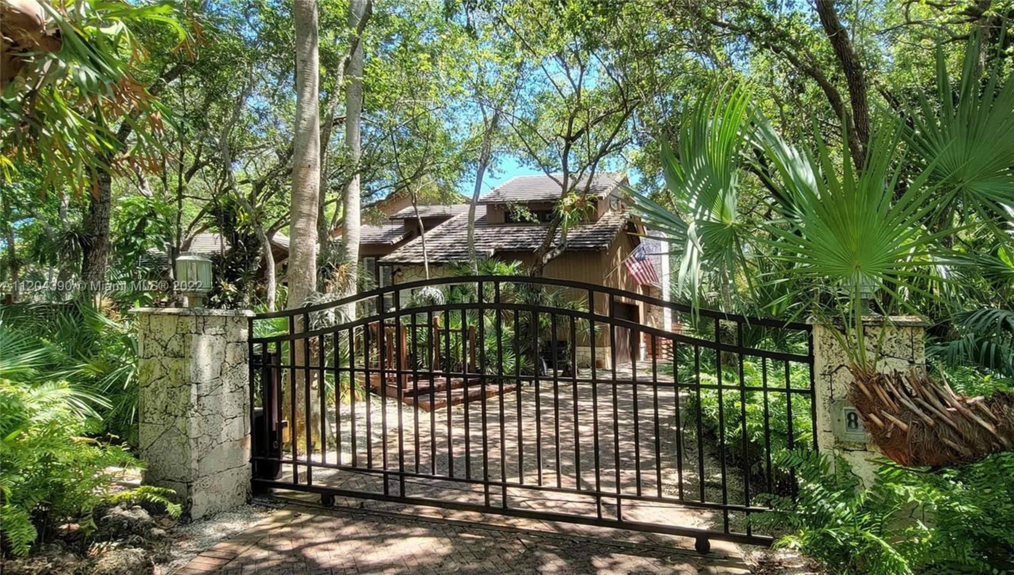 A black wrought-iron gate opens to a pathway leading to a house surrounded by trees and lush foliage.