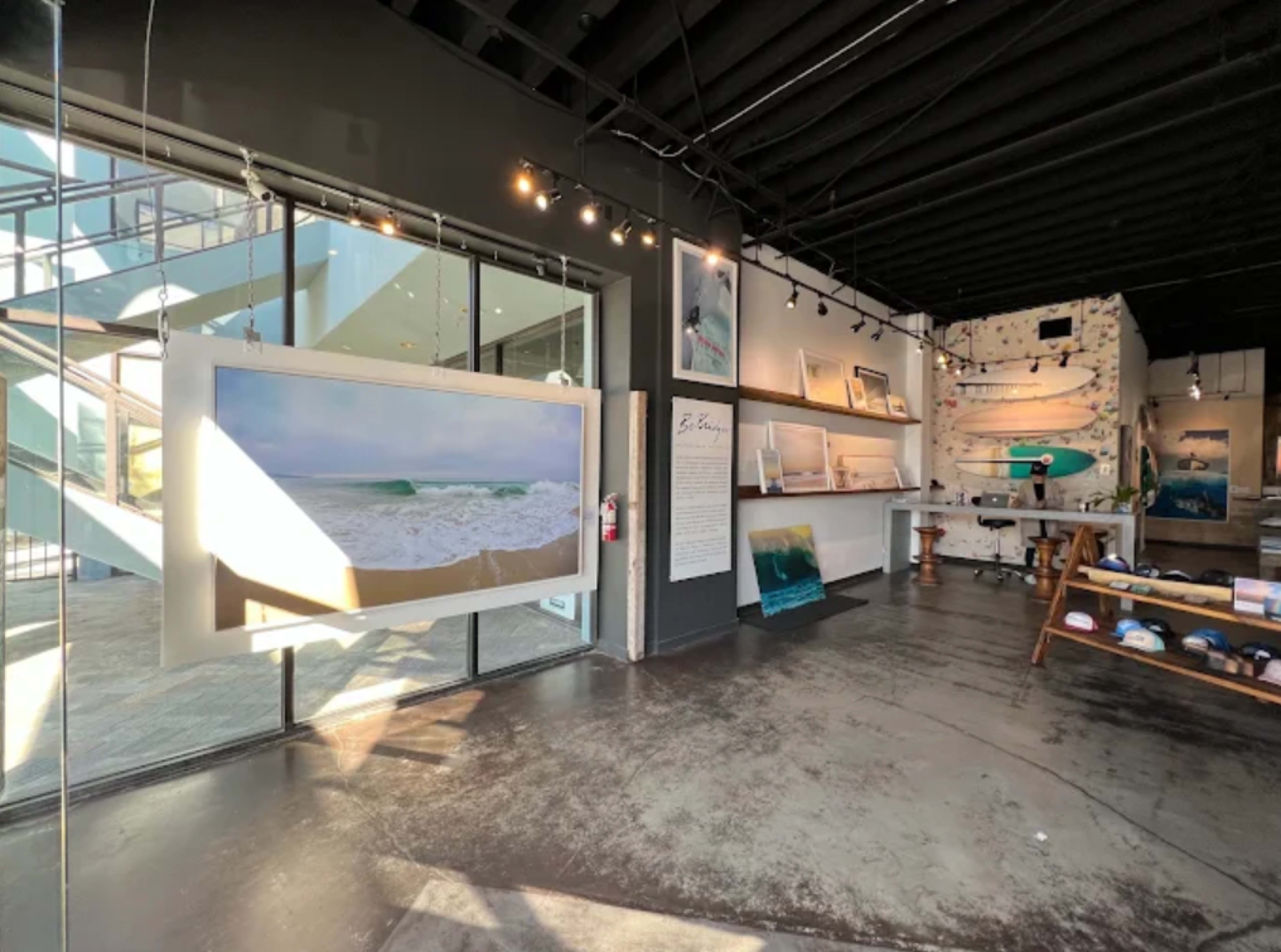 The image shows an art gallery interior with a large framed photo of ocean waves hanging prominently, surrounded by various ocean-themed artworks and merchandise displayed on shelves.