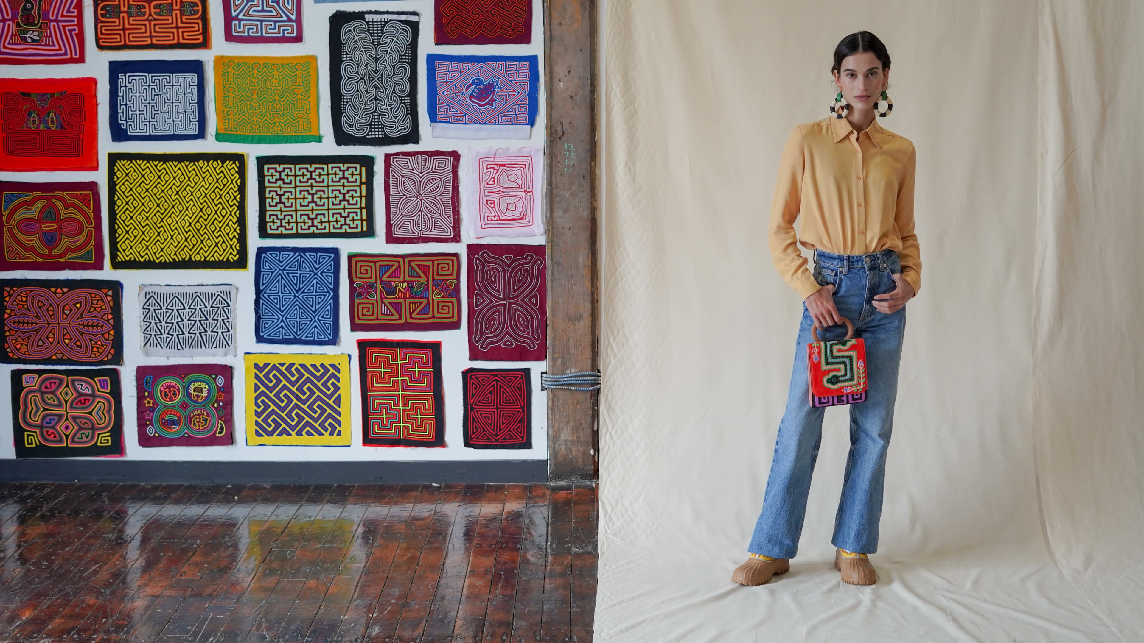 A woman stands in front of a colorful textile artwork on a wooden floor, holding a decorative bag and wearing a yellow blouse and blue jeans.
