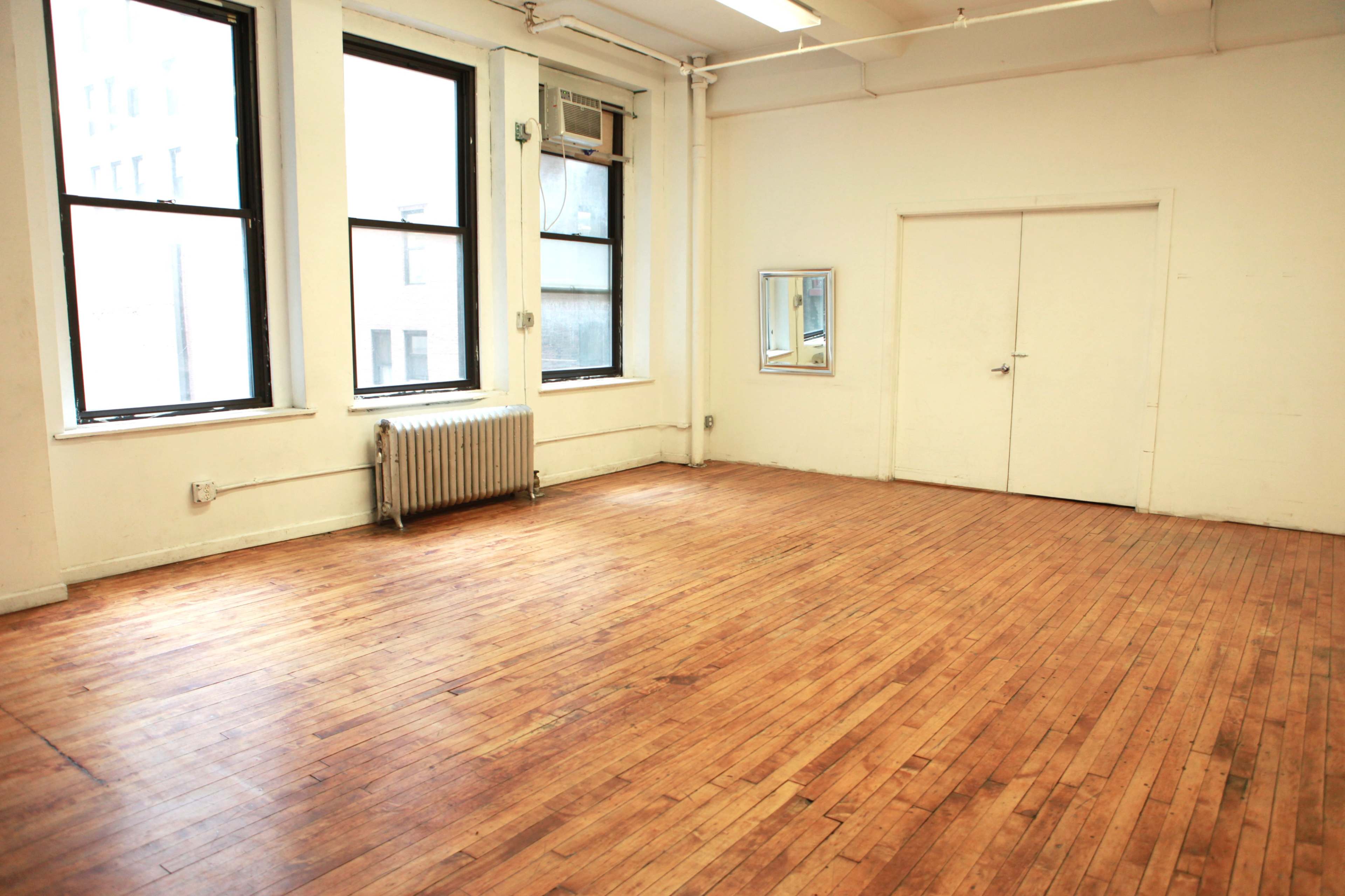 An empty room with wooden floors, large windows, and a mirrored wall reflects minimal furnishings and walls painted white.