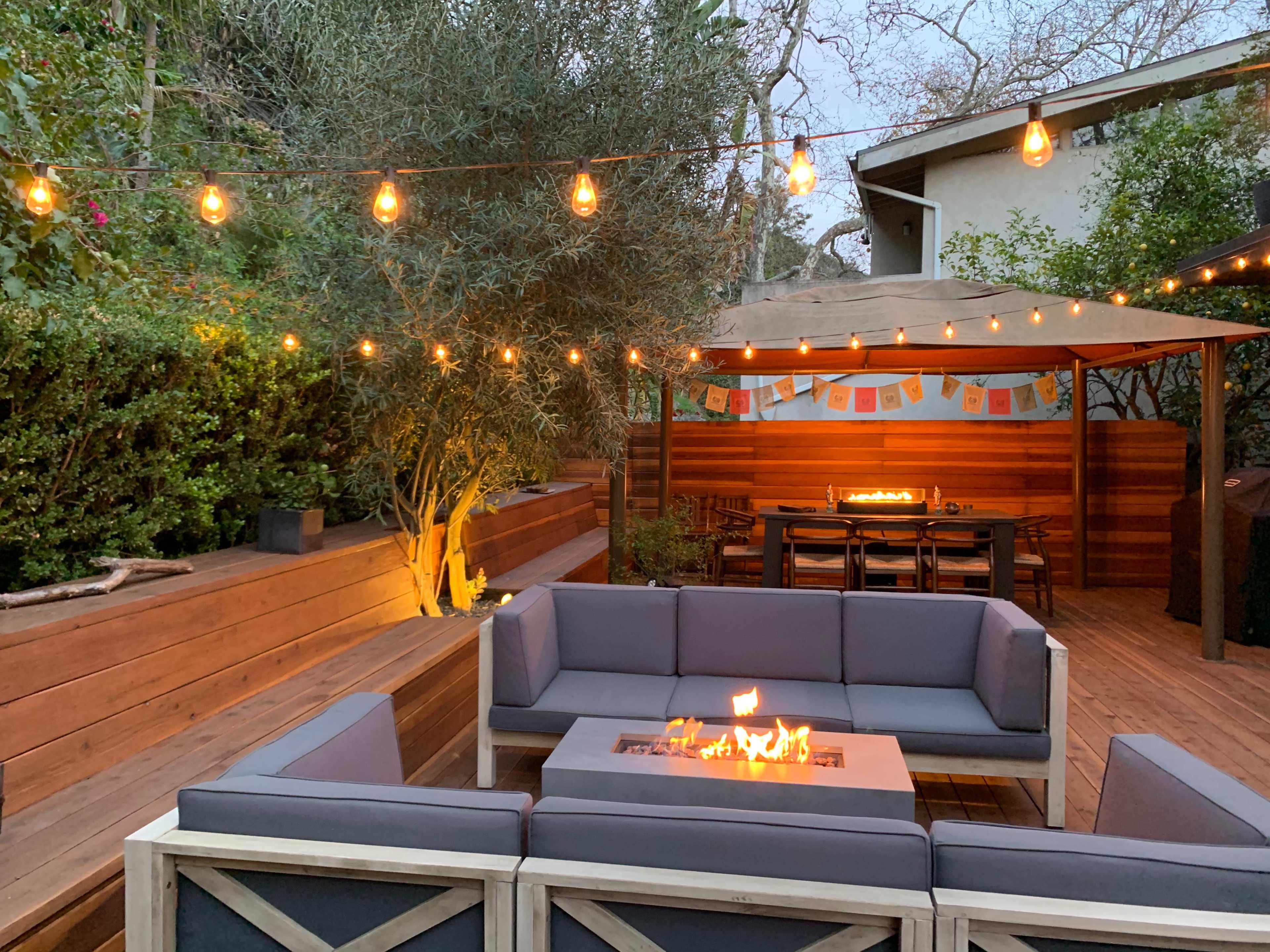 The image depicts an outdoor patio area featuring a fire pit surrounded by sectional seating, string lights overhead, and a gazebo with tables in the background.