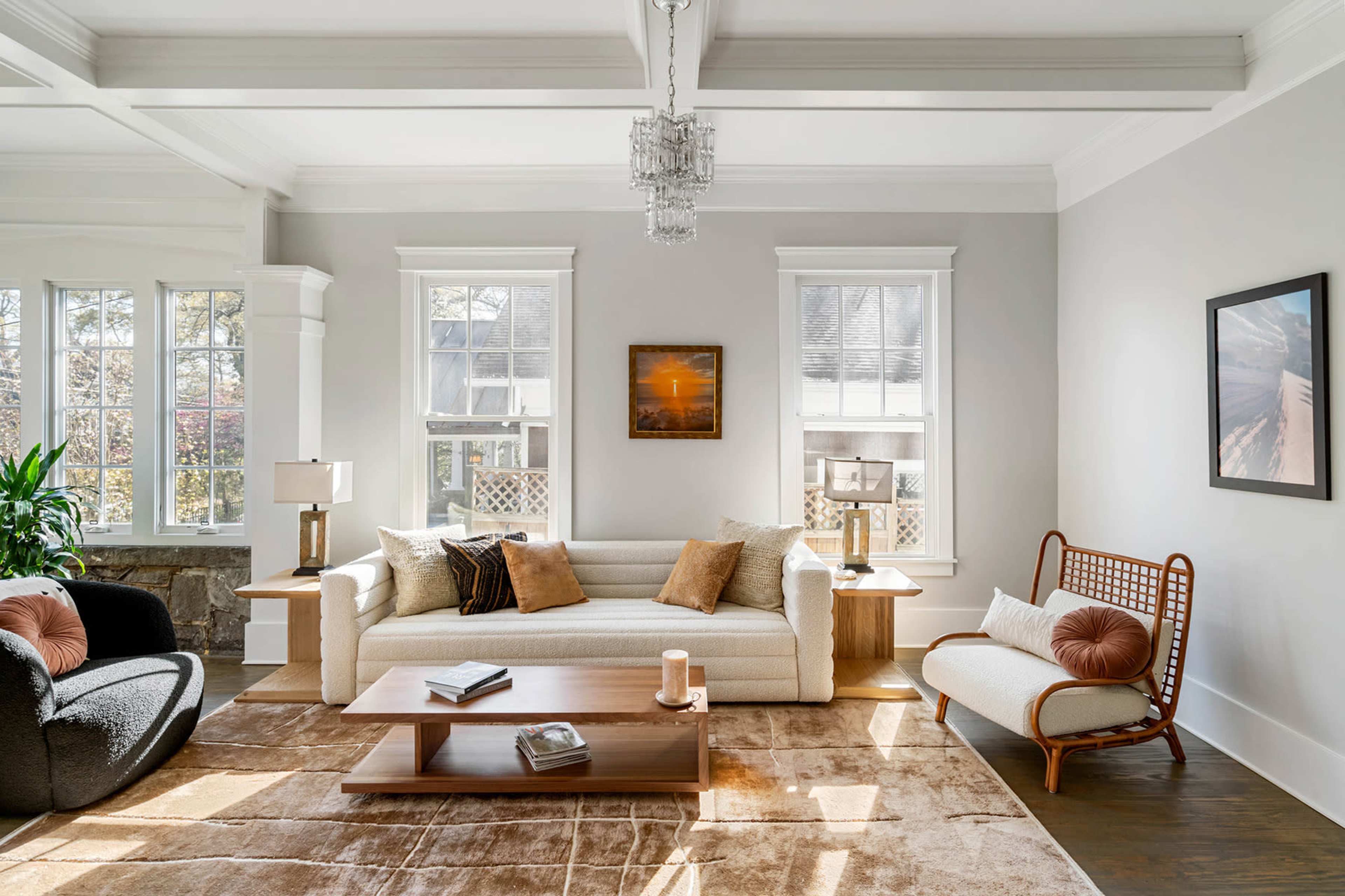 The image shows a bright living room with a white sofa, a dark armchair, a light armchair, a wooden coffee table, and large windows allowing natural light to enter.