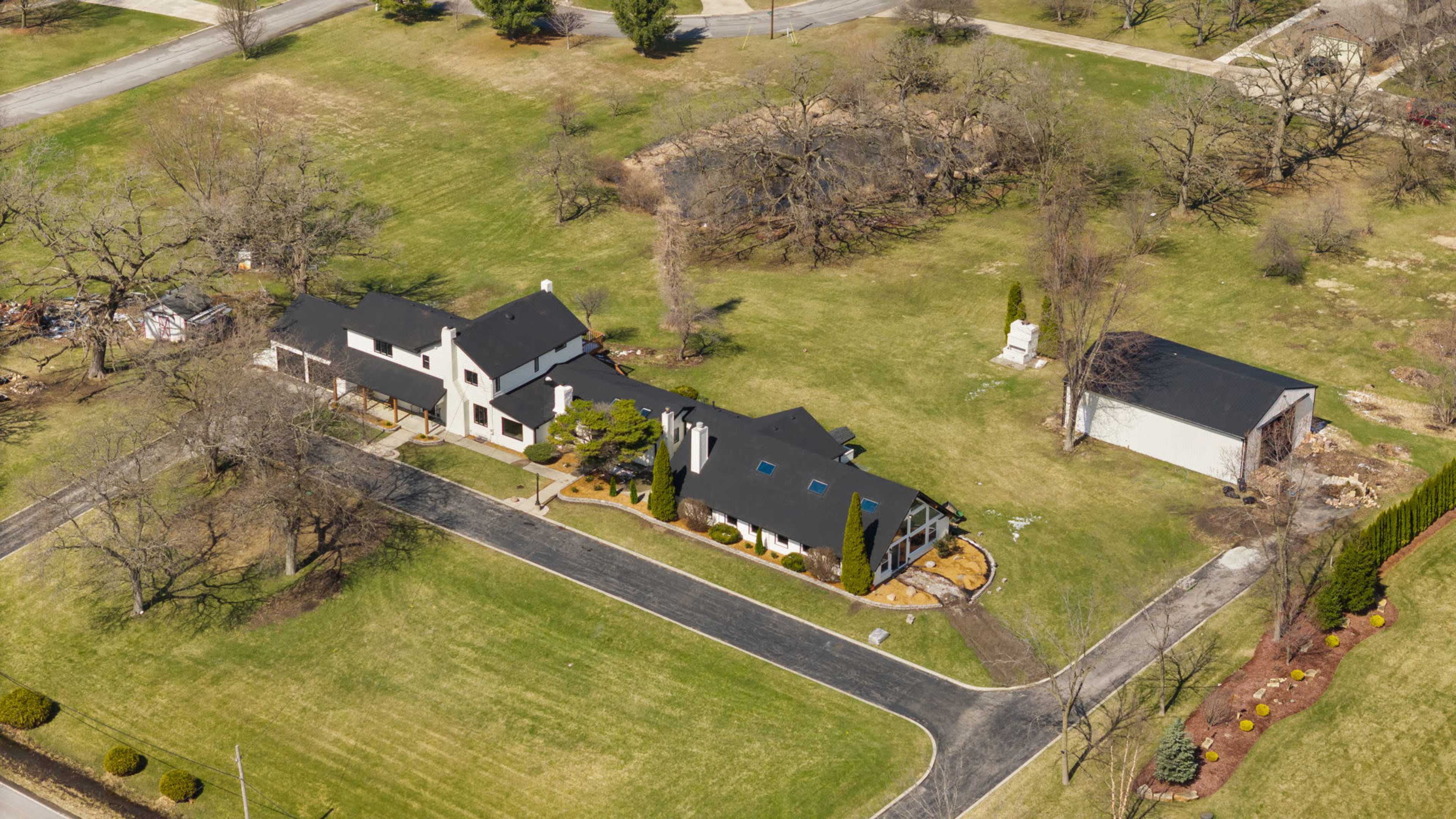 The image shows an aerial view of a spacious property featuring a large house with a dark roof, surrounded by a well-maintained lawn and several trees.