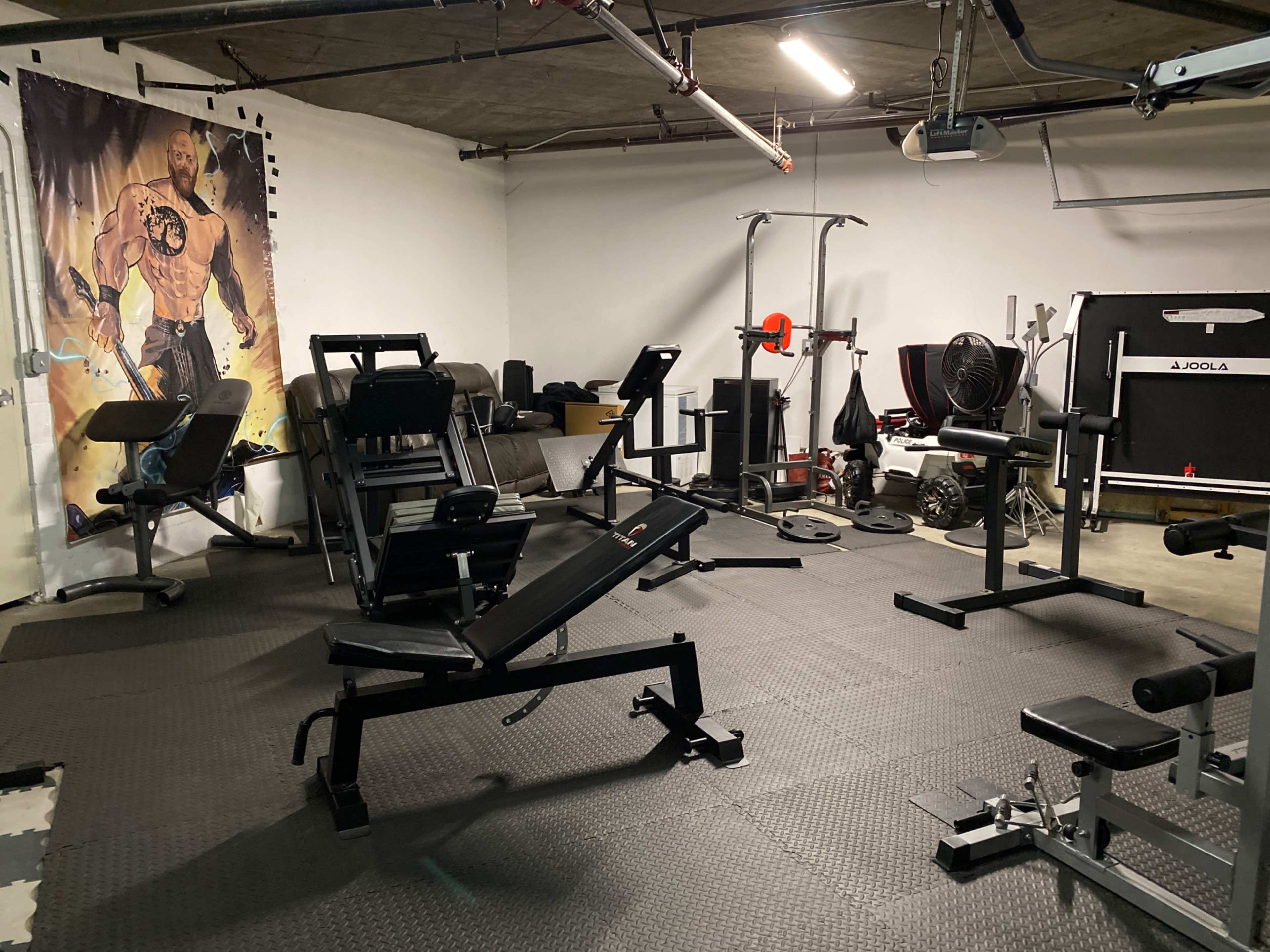 An indoor gym with various workout equipment, including benches, weights, and a wall mural of a muscular figure.