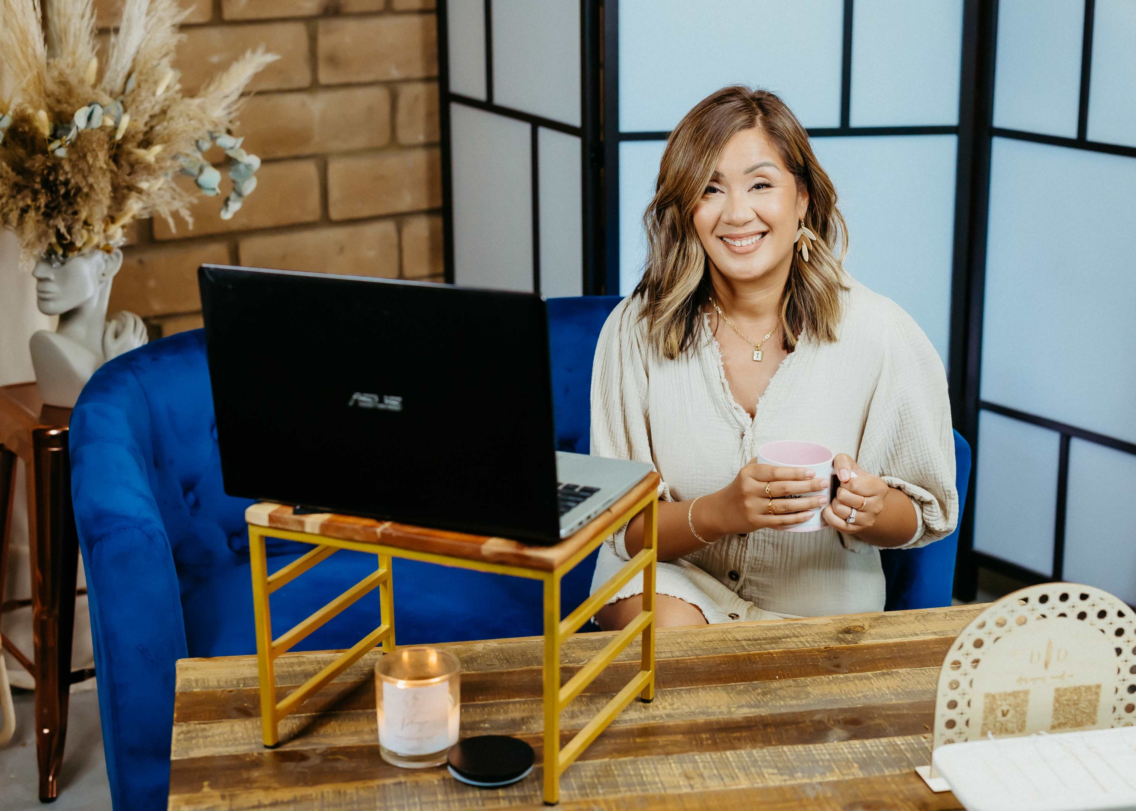 A woman sitting at a wooden table with a laptop and a cup in her hands, smiling in a well-lit room with a blue chair and decorative elements in the background.