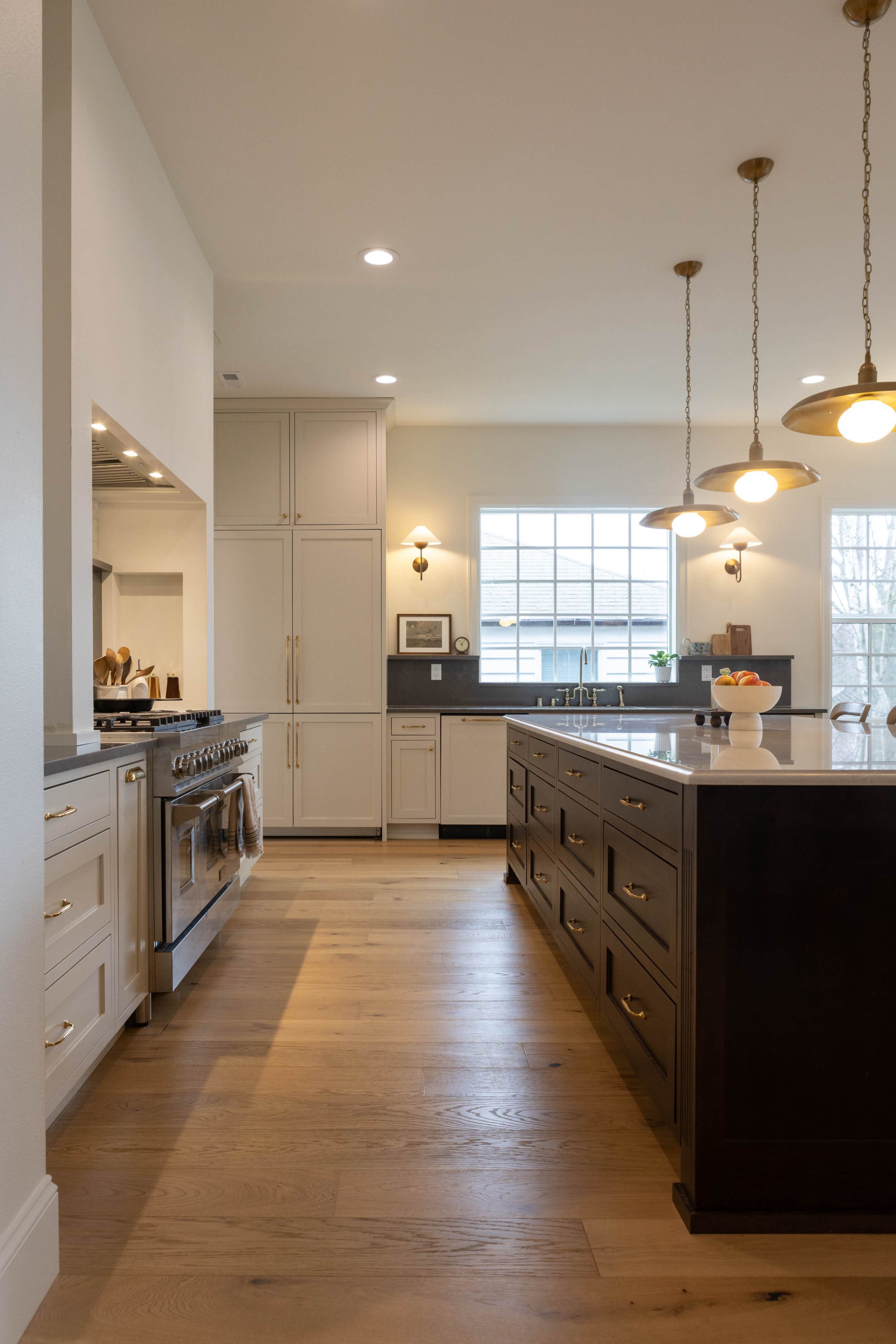 A modern kitchen features a central island with dark cabinetry, stainless steel appliances, and warm wooden flooring.