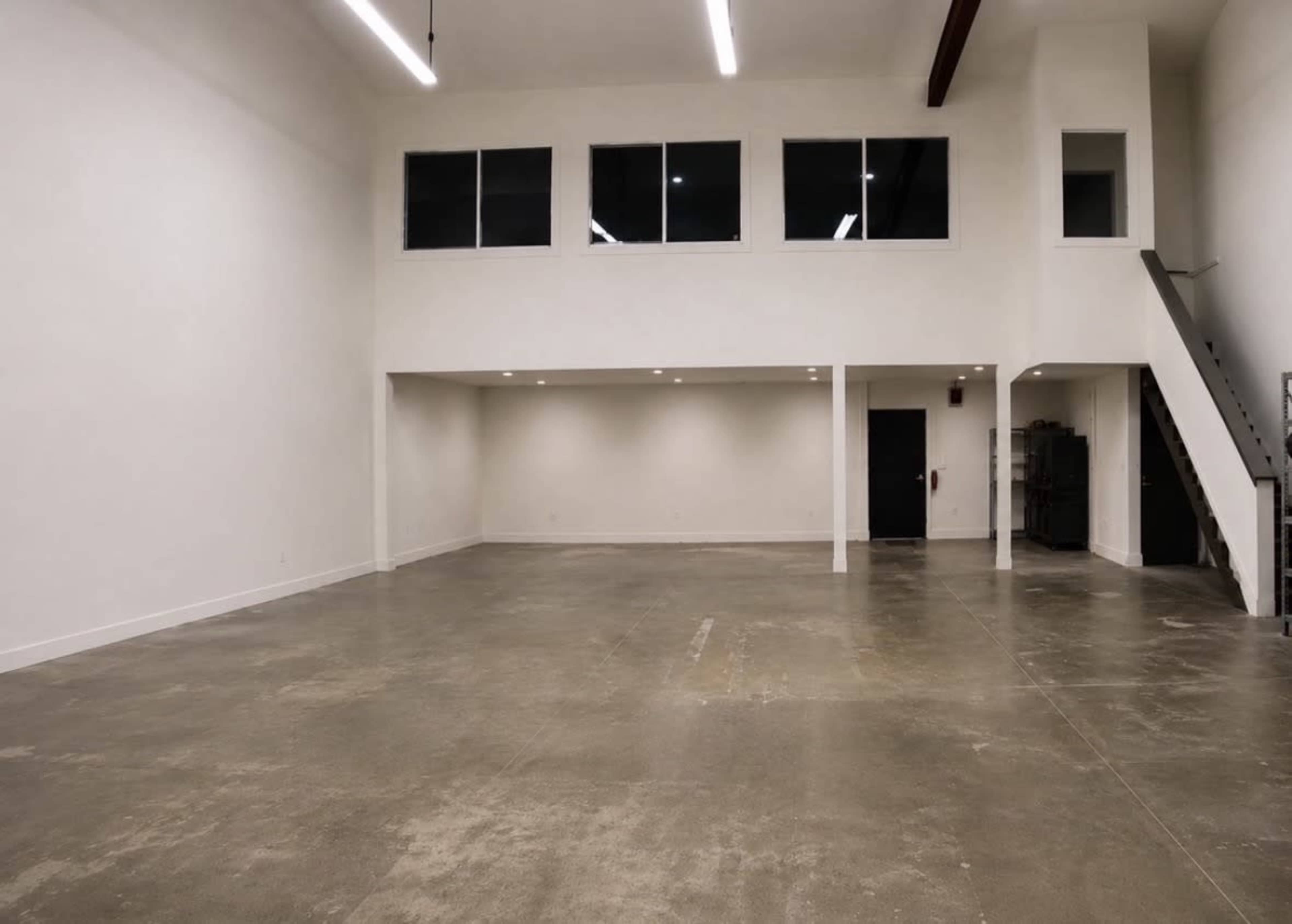 The image shows an empty, spacious interior with concrete flooring, white walls, large windows, and a staircase leading to a raised area.