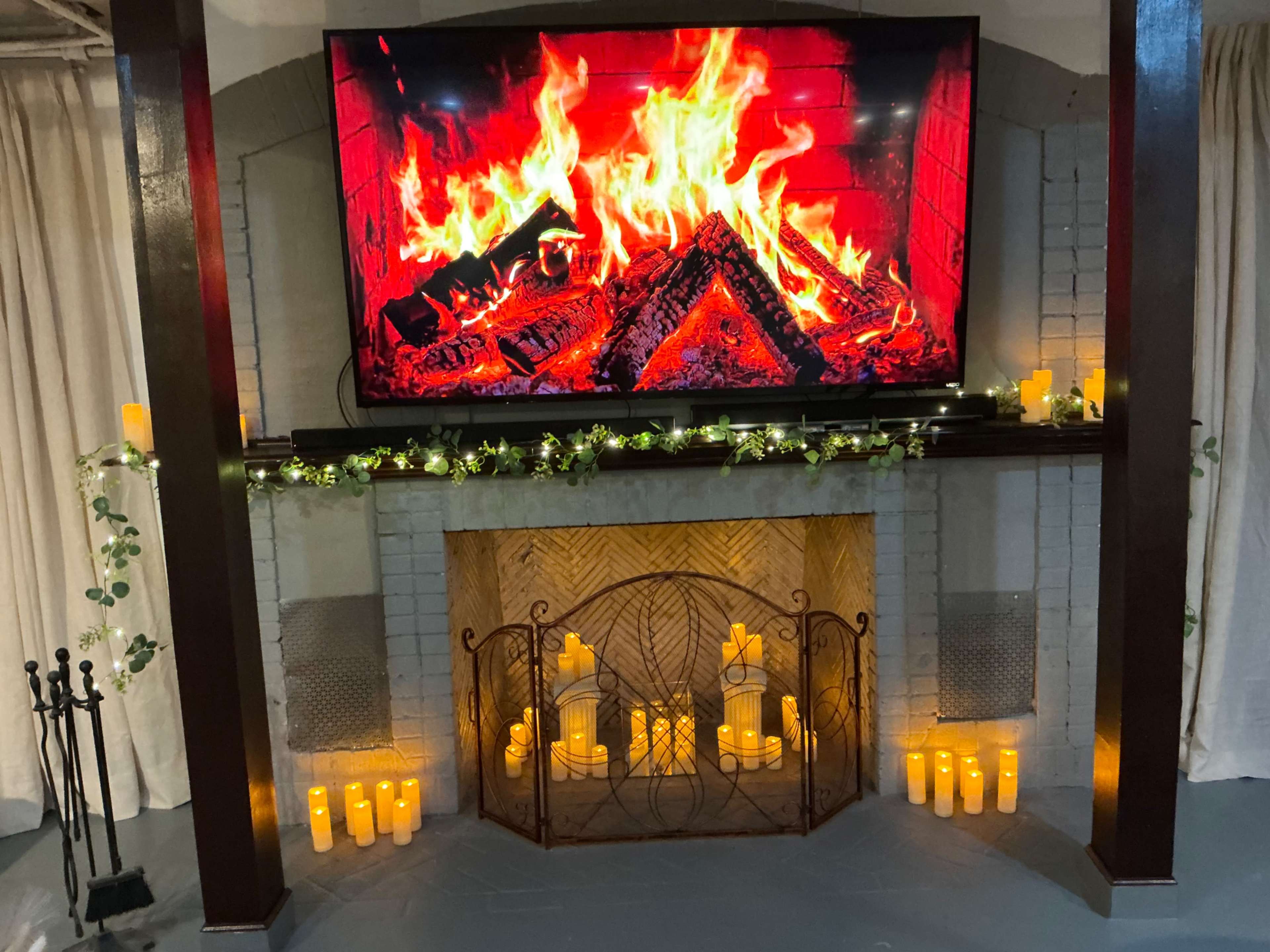The image shows a decorative fireplace setup featuring a large TV displaying a fire graphic, surrounded by candles and greenery.