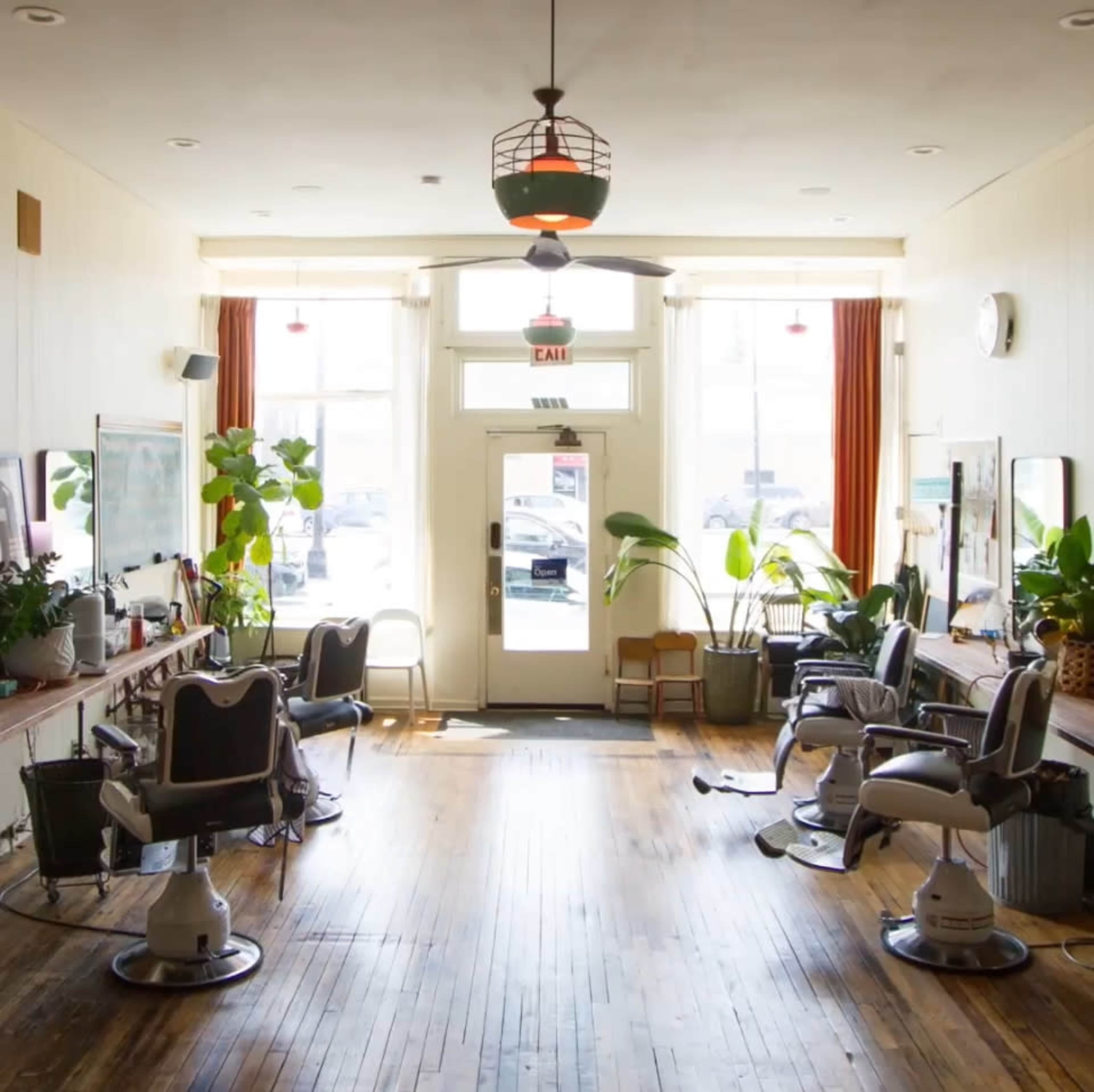 The interior of a hair salon features several barber chairs and large windows with plants lining the entrance.