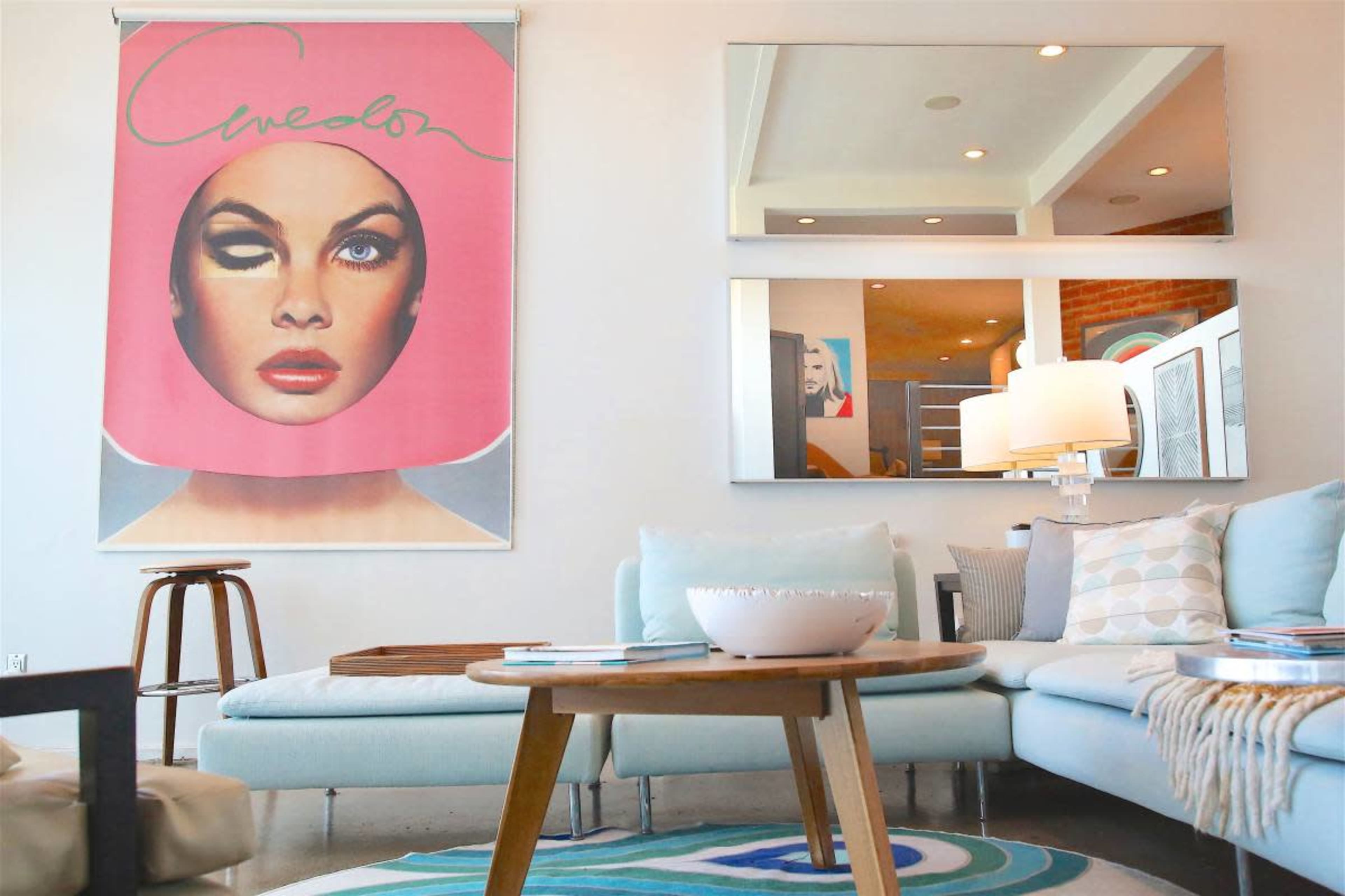 The image features a modern living room with a large pink wall hanging of a woman's face, a light blue sectional sofa, a wooden coffee table, and mirrors reflecting the space.