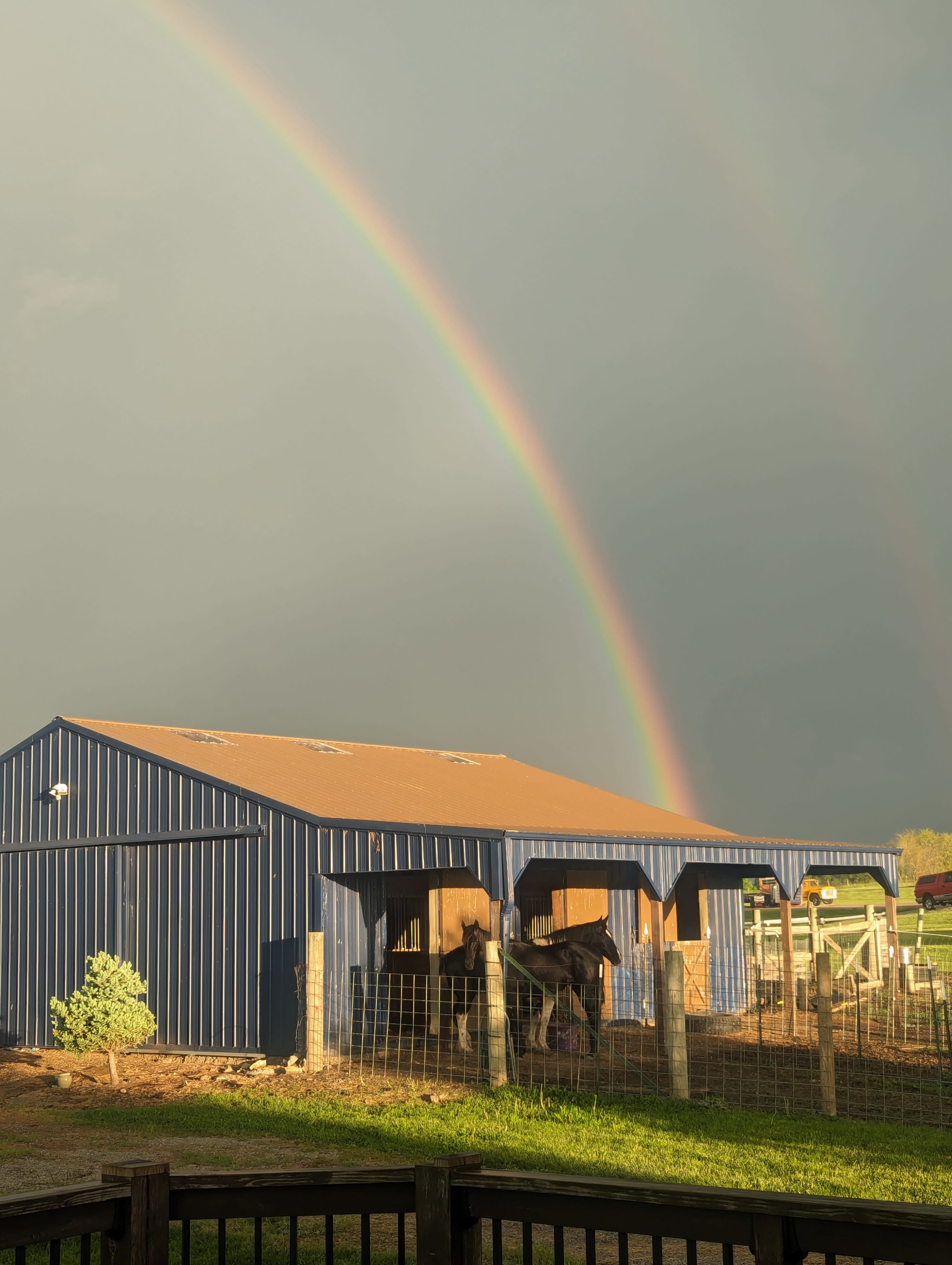 A blue barn stands under a double rainbow, with a horse visible in a nearby pen.