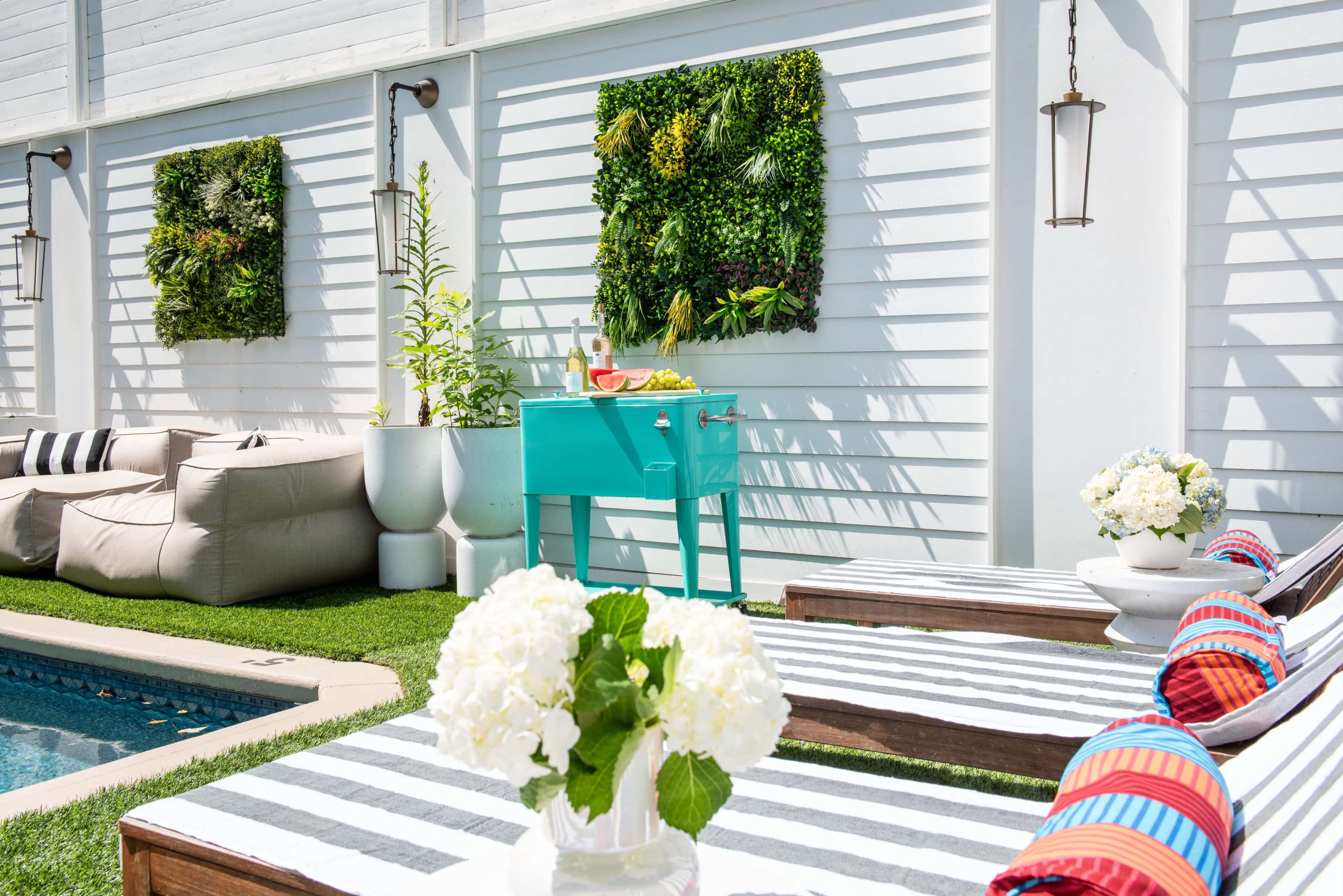 The scene features a modern outdoor space with lounge chairs, a turquoise grill, potted plants, and wall-mounted greenery beside a pool.