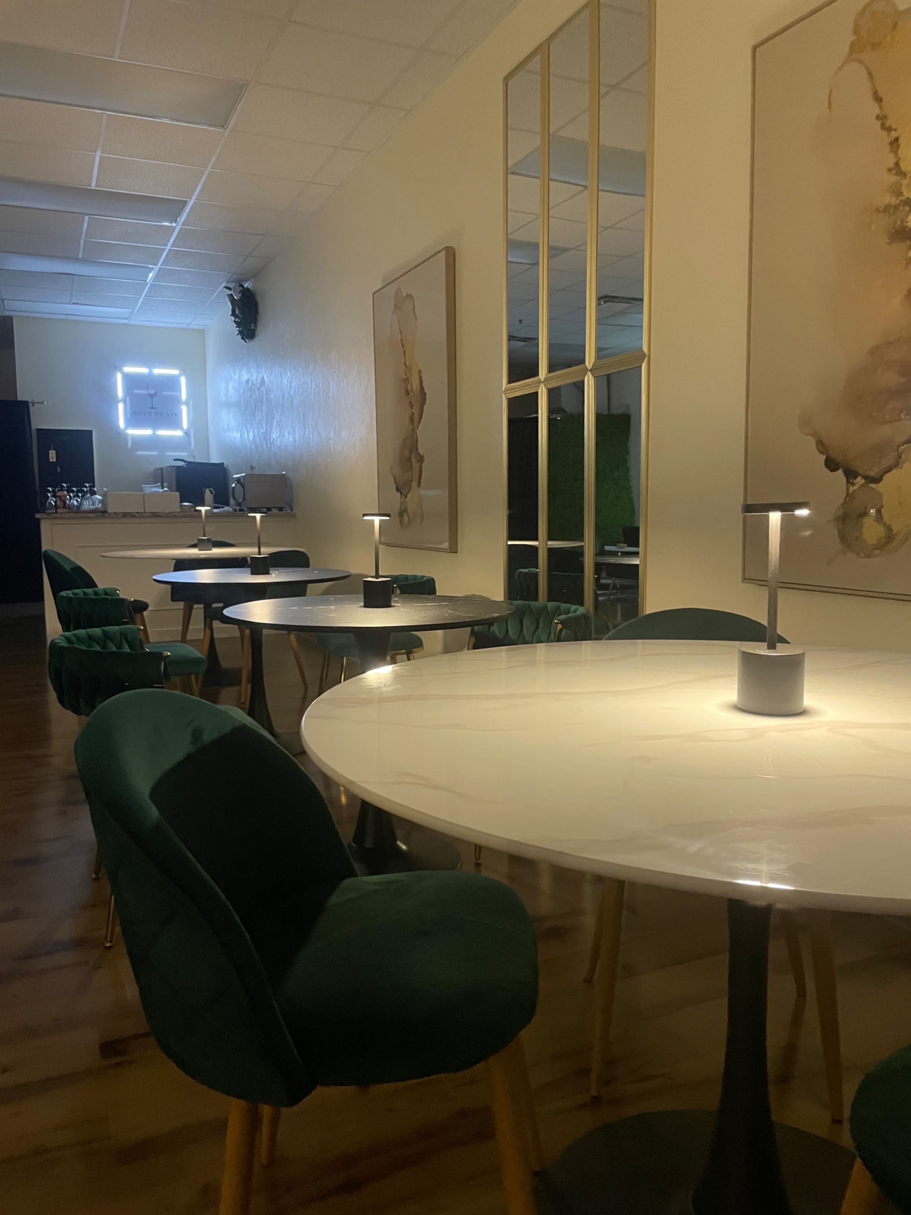 The image shows a modern, stylish interior of a café or workspace featuring round marble-topped tables with green upholstered chairs and decorative wall art.