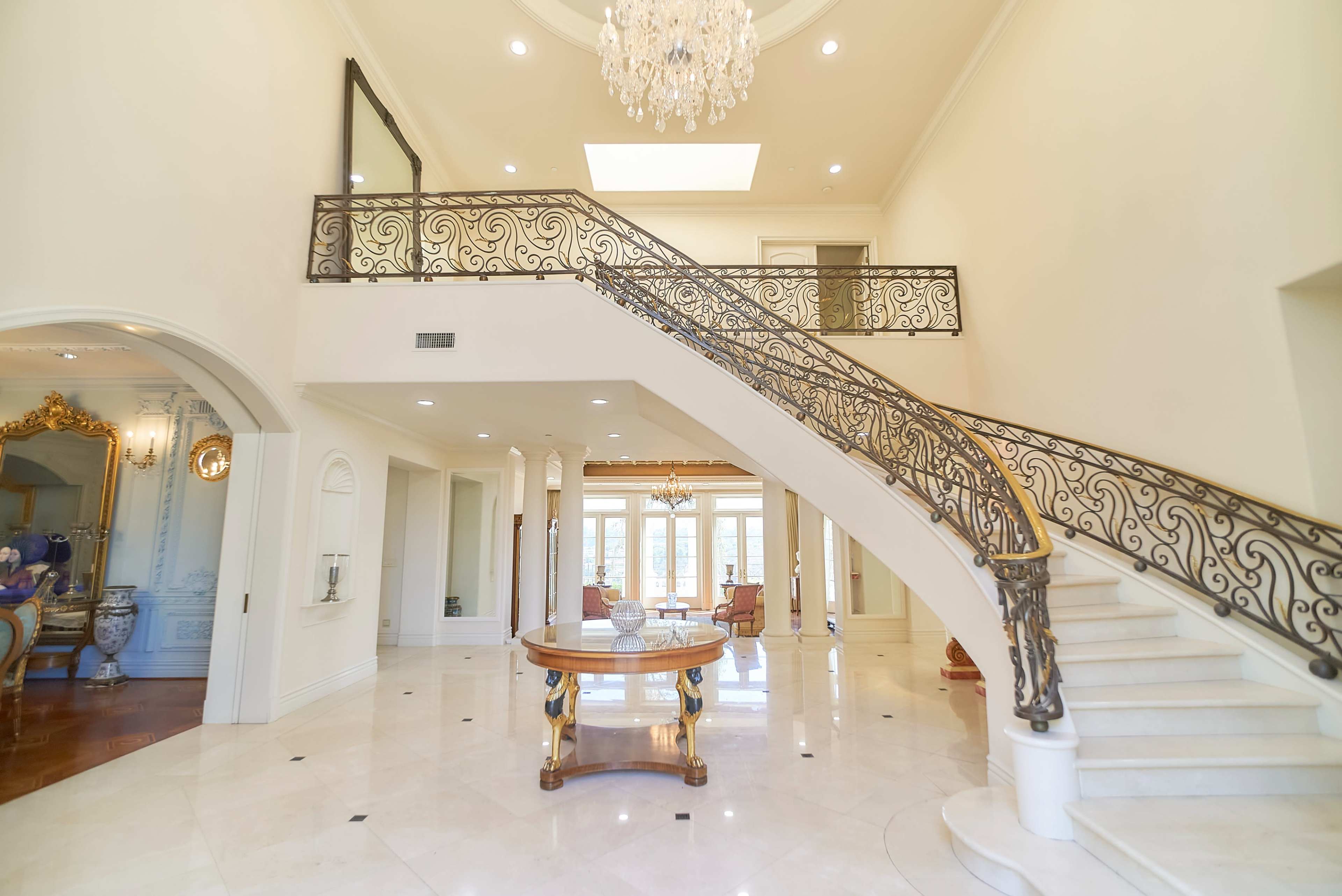 A spacious foyer with a grand staircase featuring ornate railings, a chandelier overhead, and marble flooring.