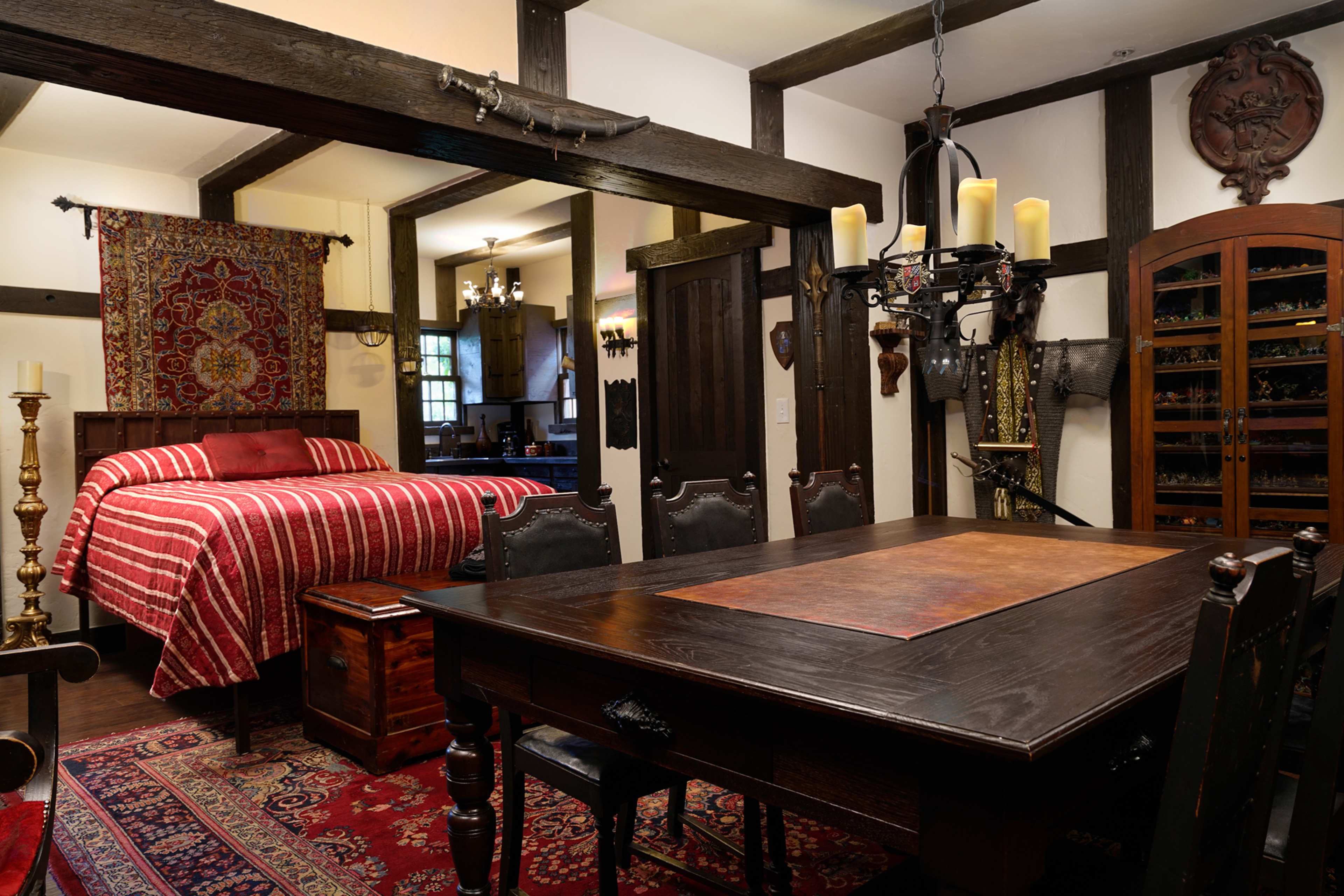 The image shows a cozy room featuring a red-striped bed, a rustic dining table, and medieval-themed decor, including armor and a chandelier.