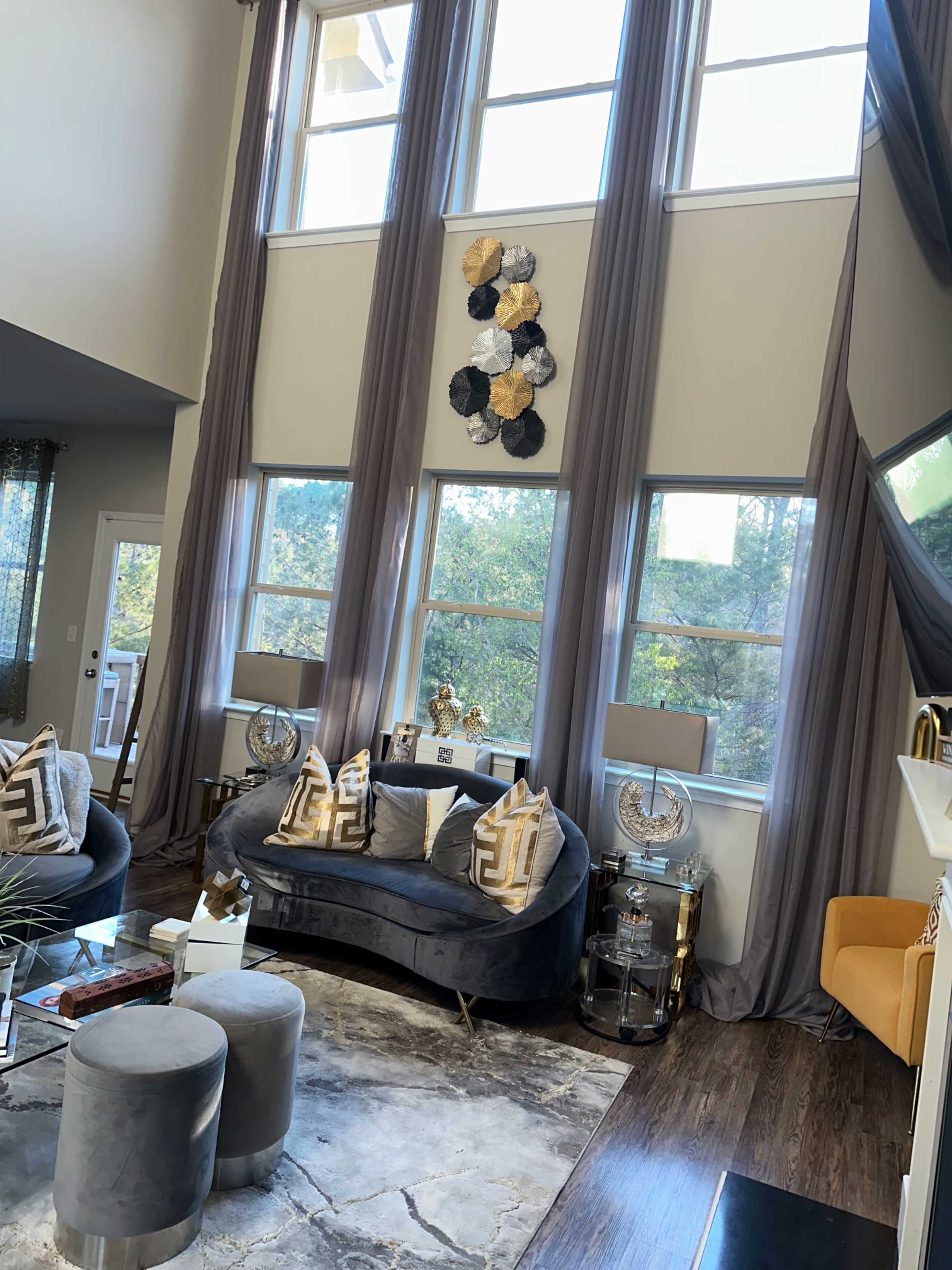 A modern living room with tall windows, gray curtains, a plush sofa, decorative pillows, and a coffee table.