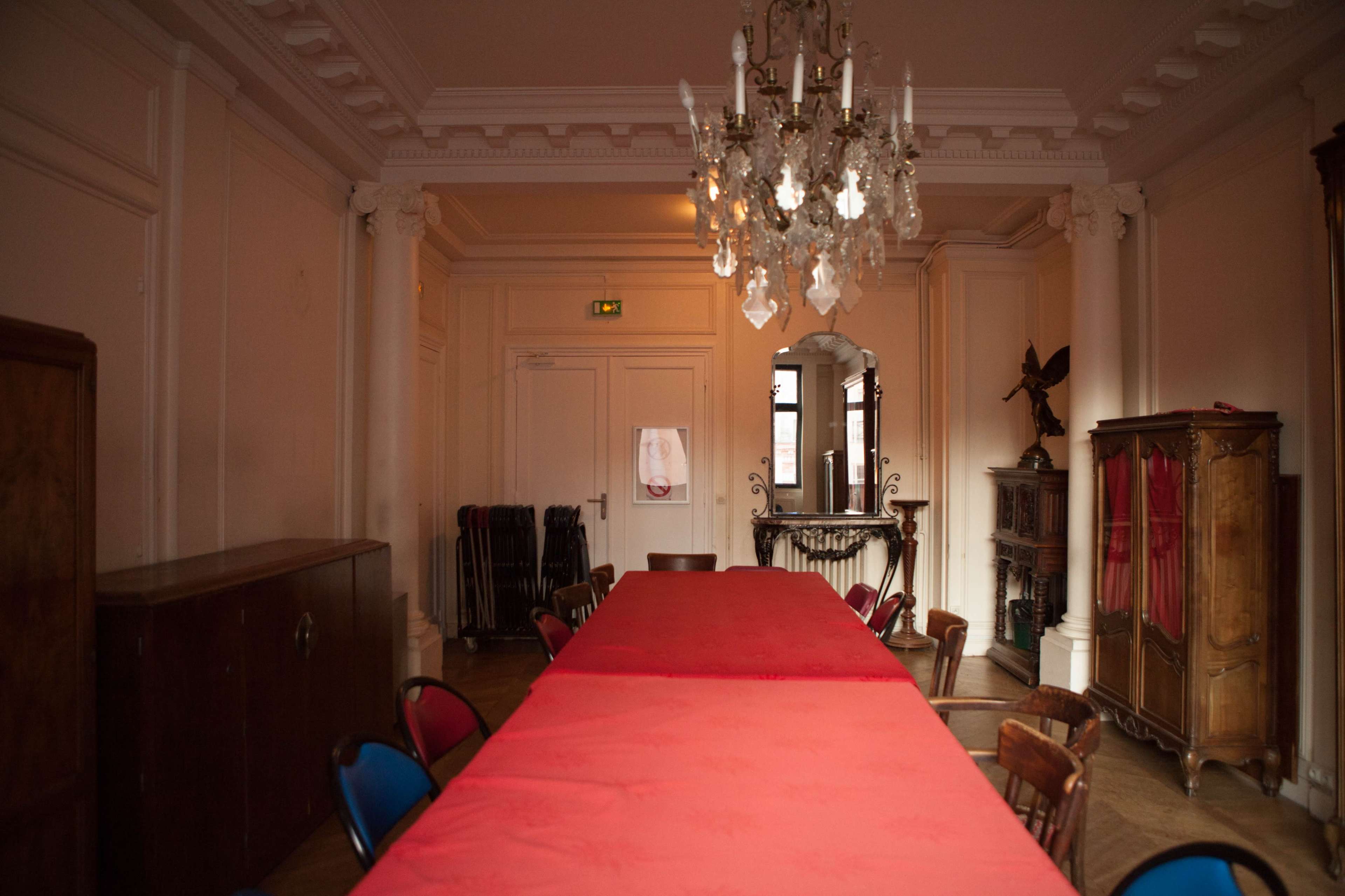 The image shows a dimly lit meeting room with a long red table, surrounded by chairs, and a chandelier hanging from the ceiling.