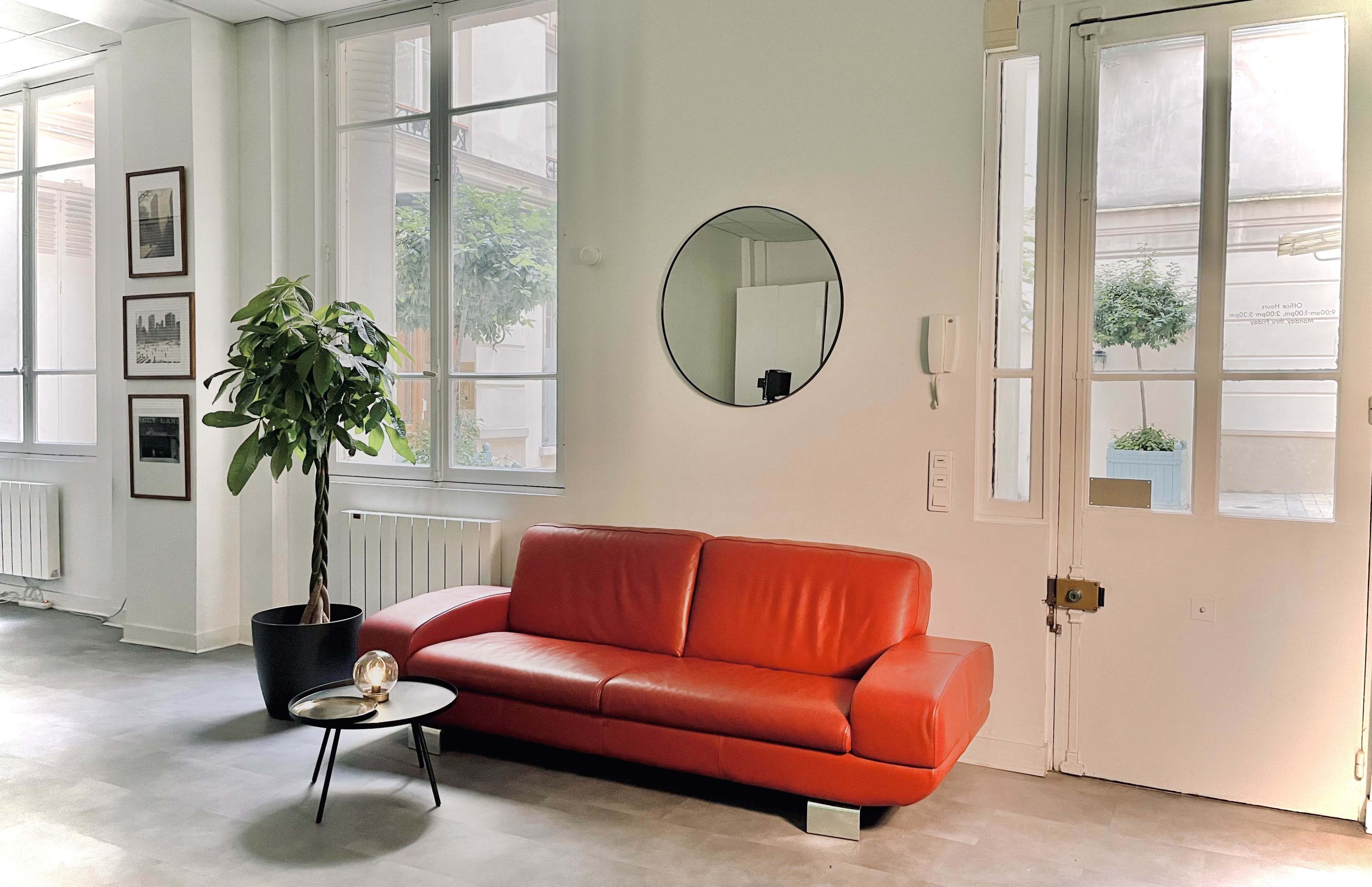 The image shows a modern, minimalist interior featuring a red sofa, a small round table, a potted plant, and large windows that let in natural light.