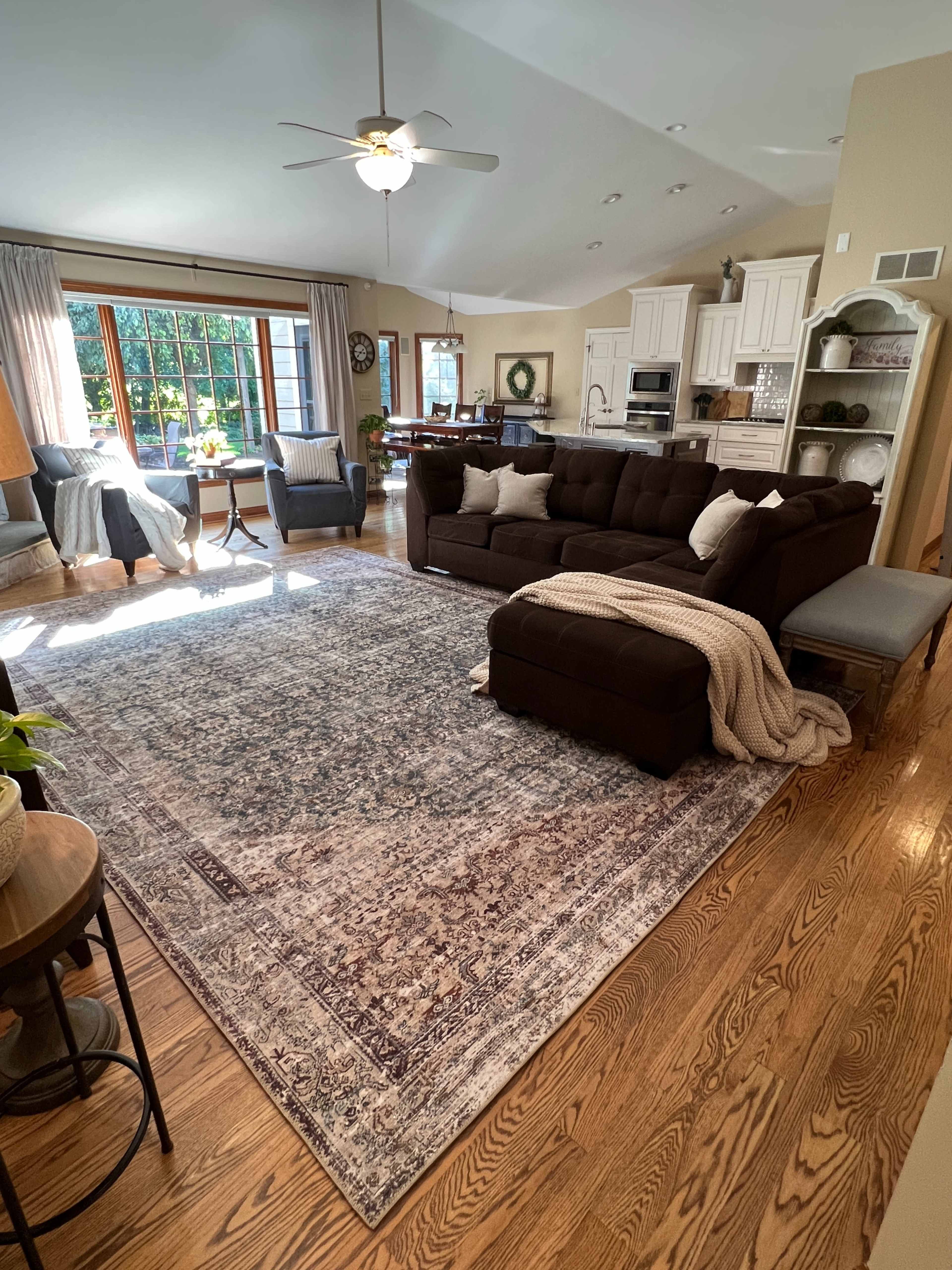 The image shows a spacious living room with a large dark sectional couch, a decorative rug, and a bright window overlooking a garden.