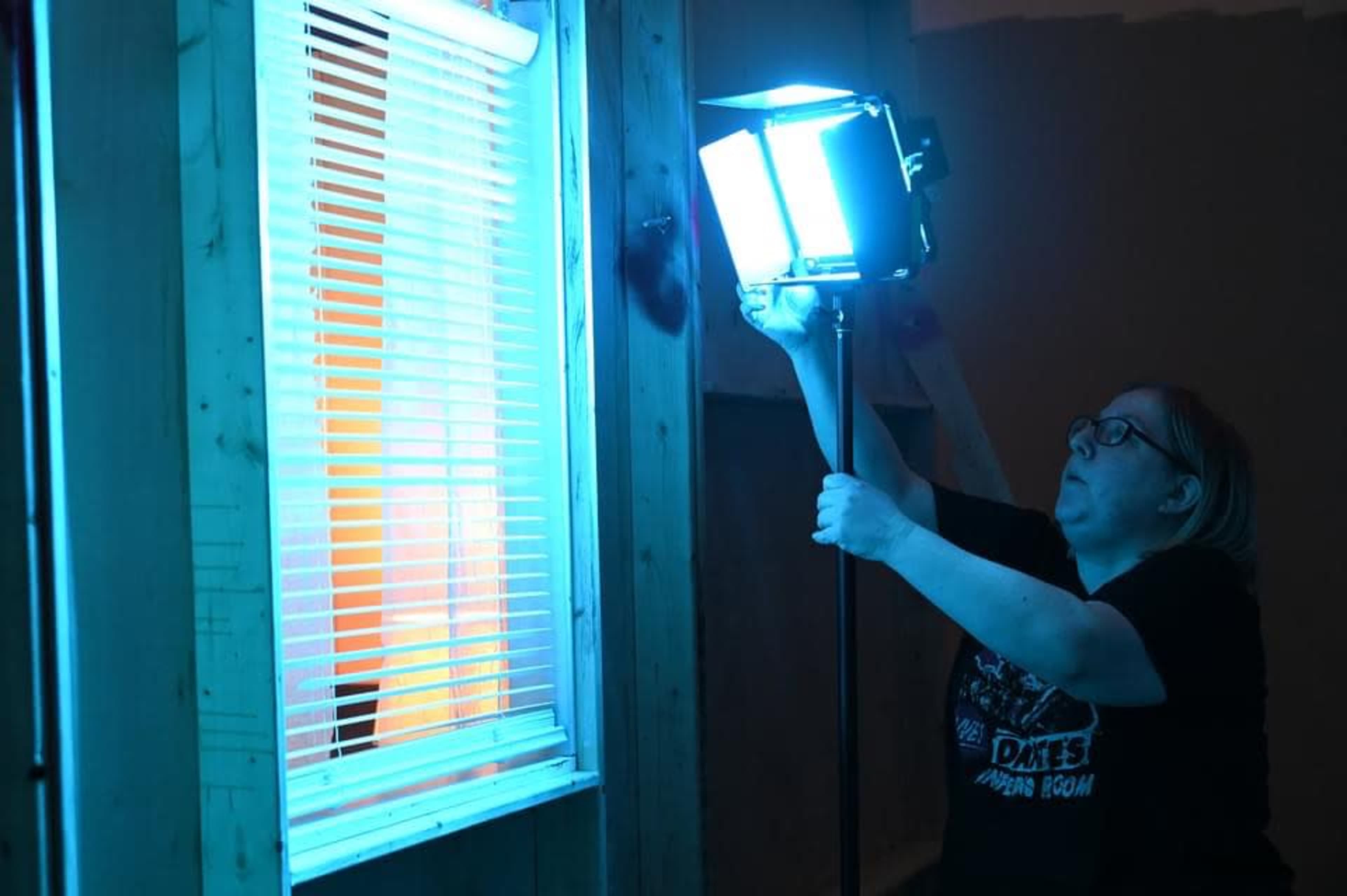 A woman adjusts a bright lighting setup near a window with blinds in a dimly lit room.