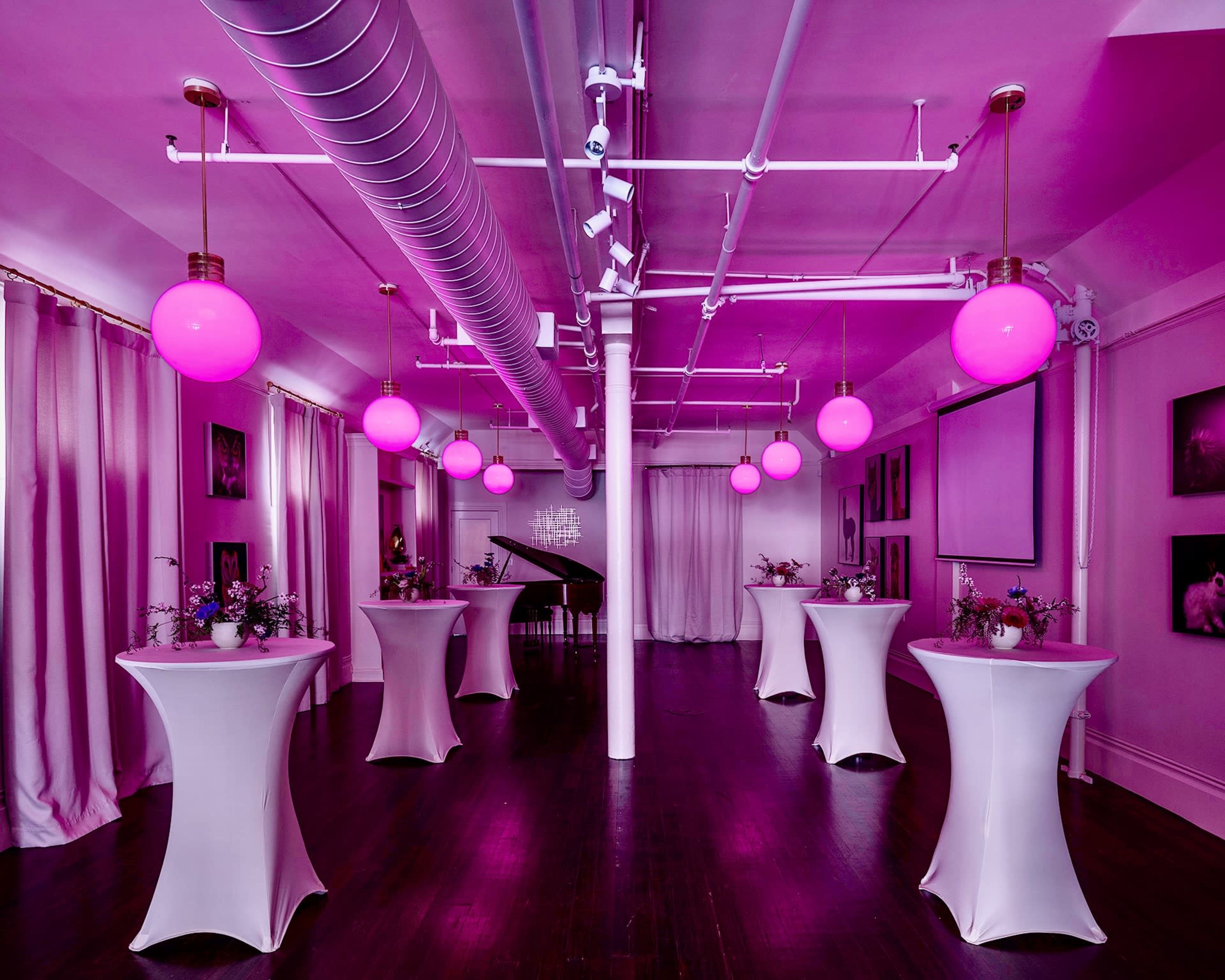 A spacious event room features several tall tables adorned with flowers and illuminated by pink globe pendant lights.