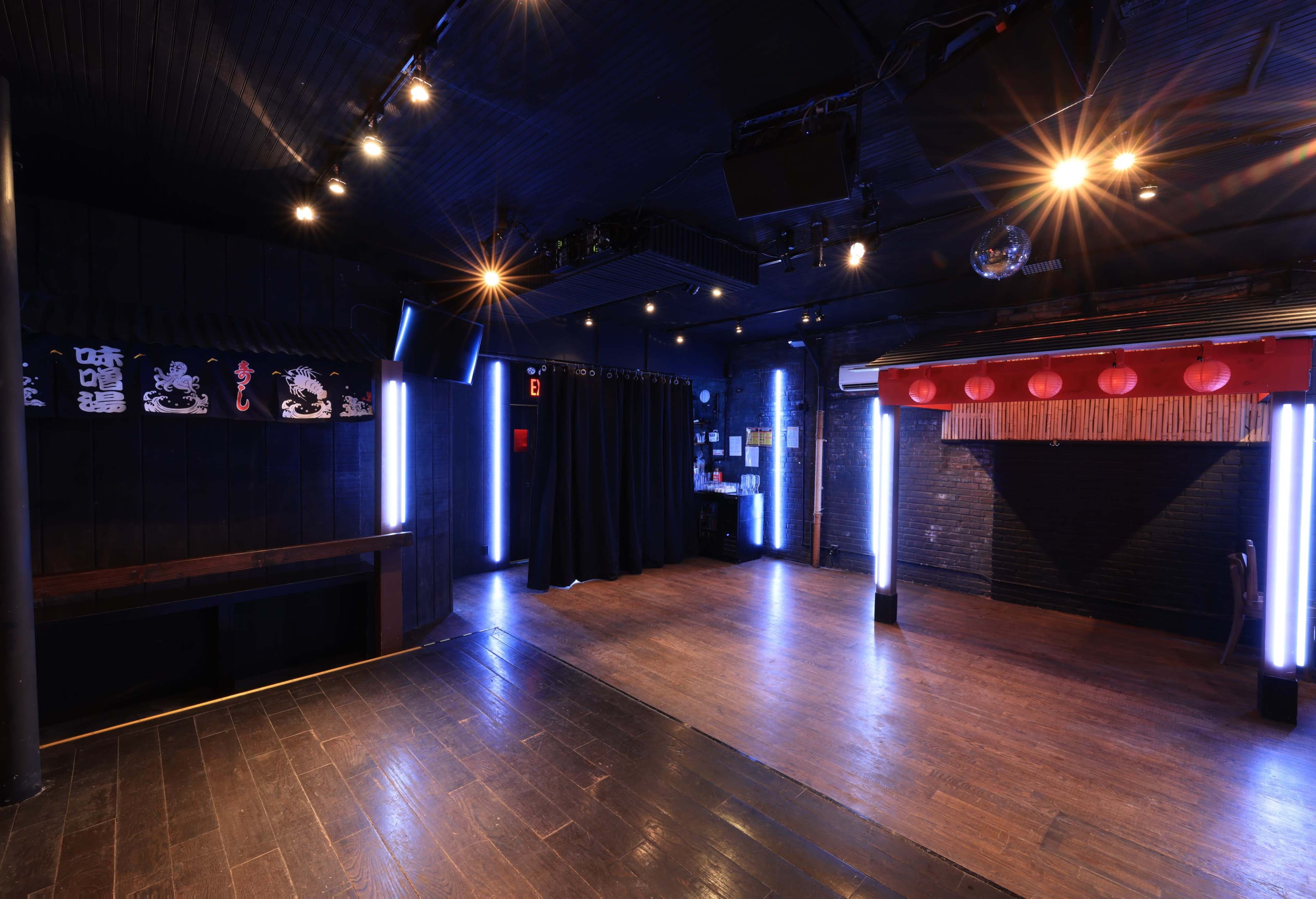 A dark, spacious interior of a performance venue with wooden flooring, illuminated by various lighting fixtures, including neon lights and spotlights.