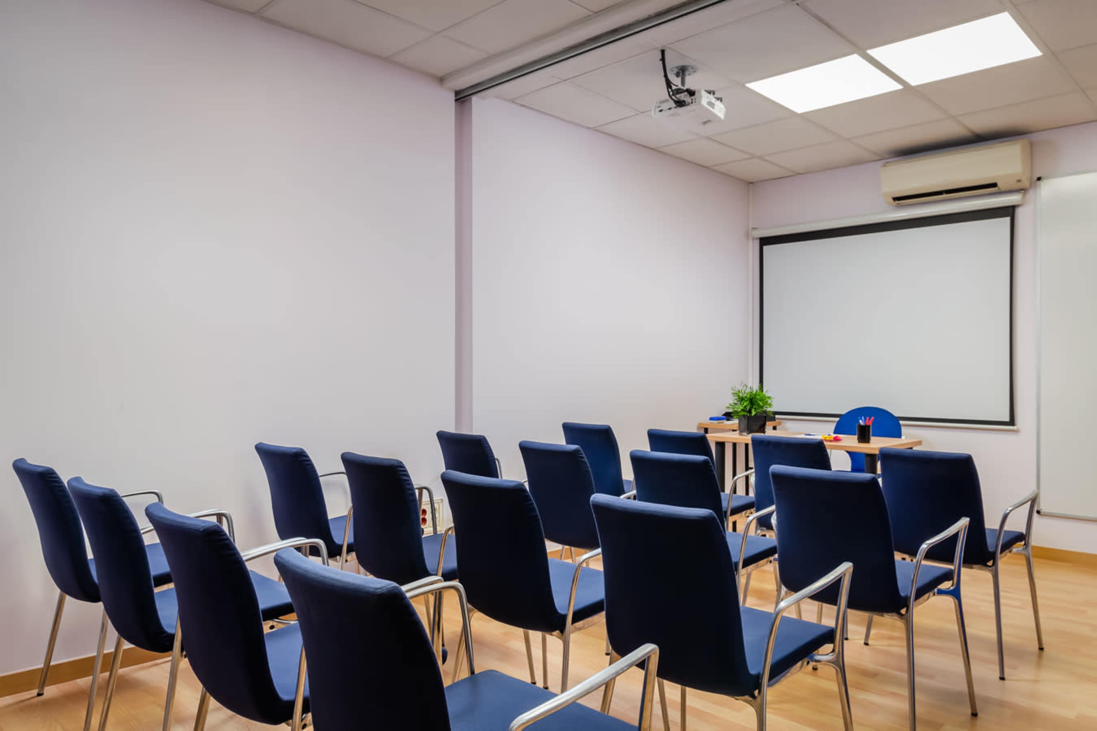 A conference room features rows of blue chairs facing a presentation area with a table, projector, and a plant.